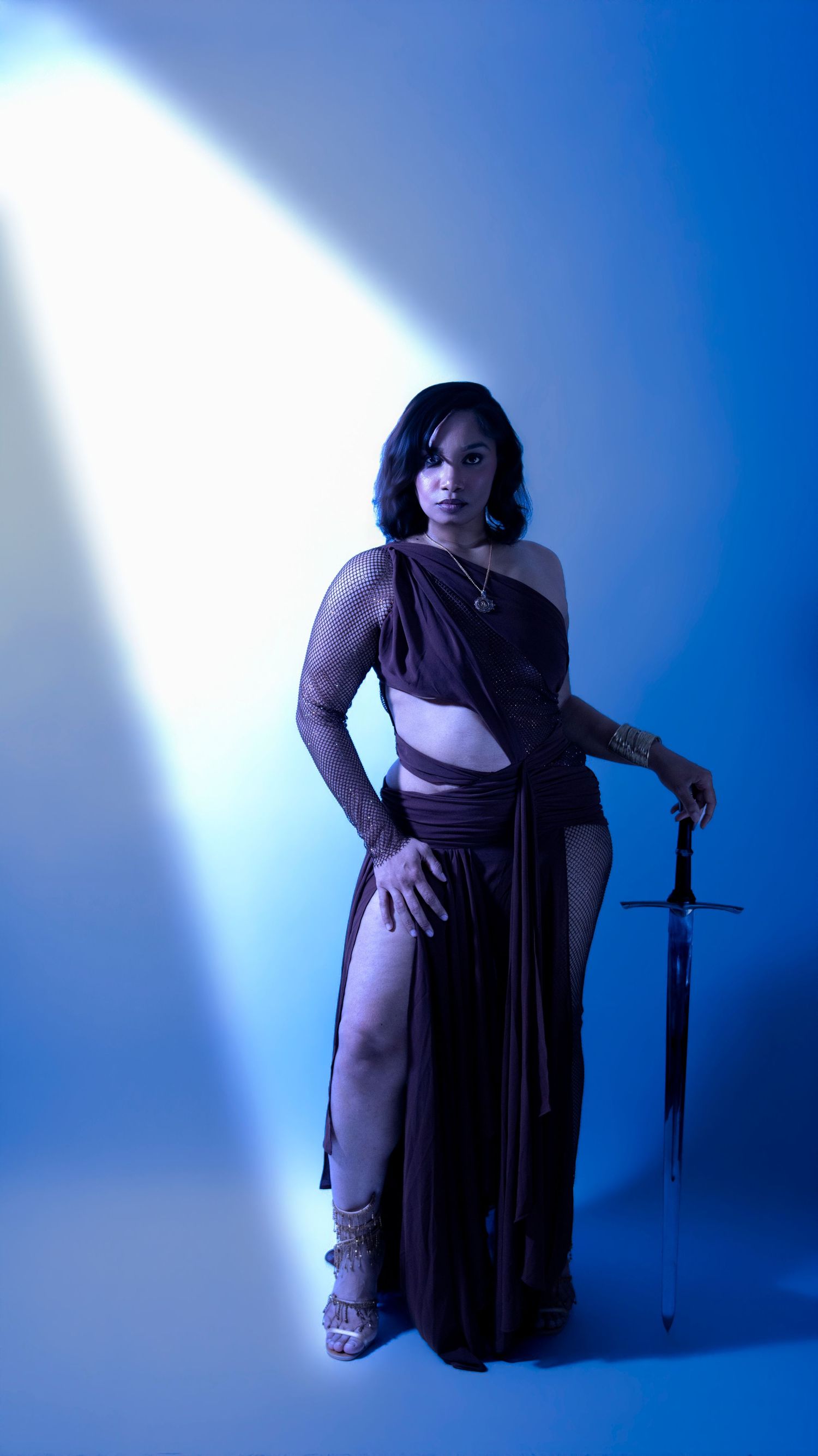 Woman in elegant brown one-shoulder cutout dress holding sword in blue-lit studio setting.