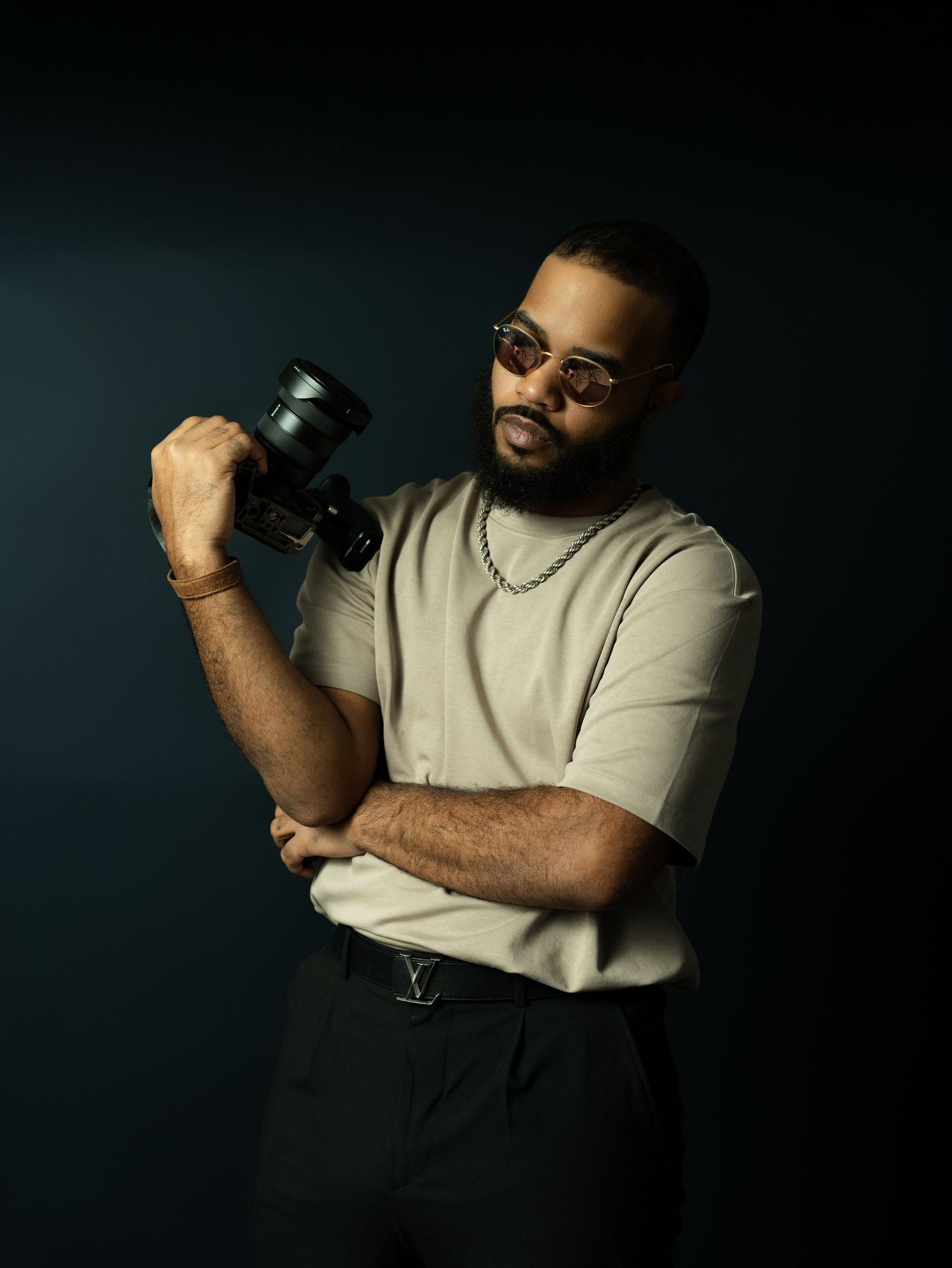 Stylish Black man with sunglasses holds camera while posing against dark background in beige tee.