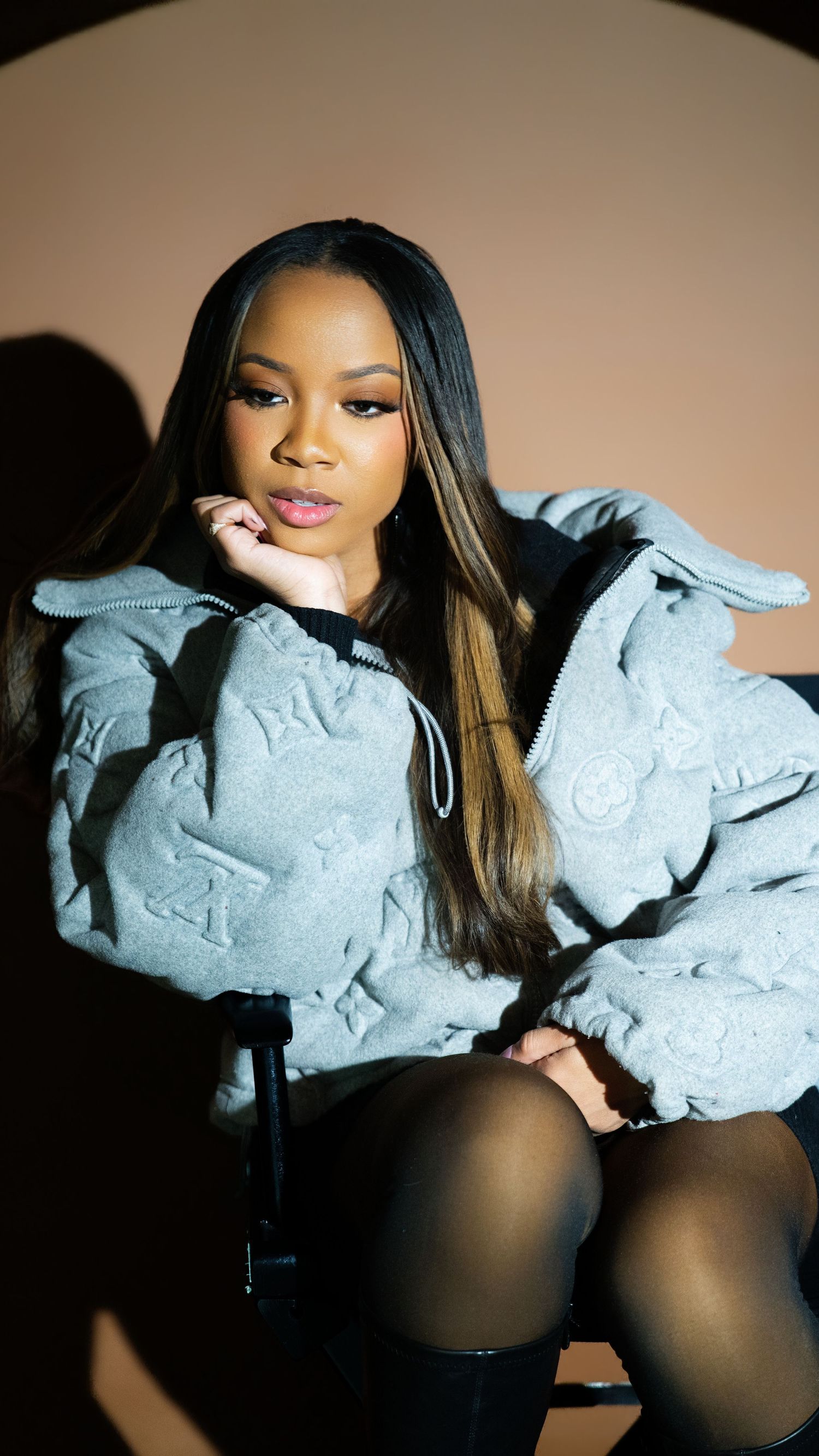 Young woman in oversized gray LV monogram puffer jacket resting chin on hand in studio.