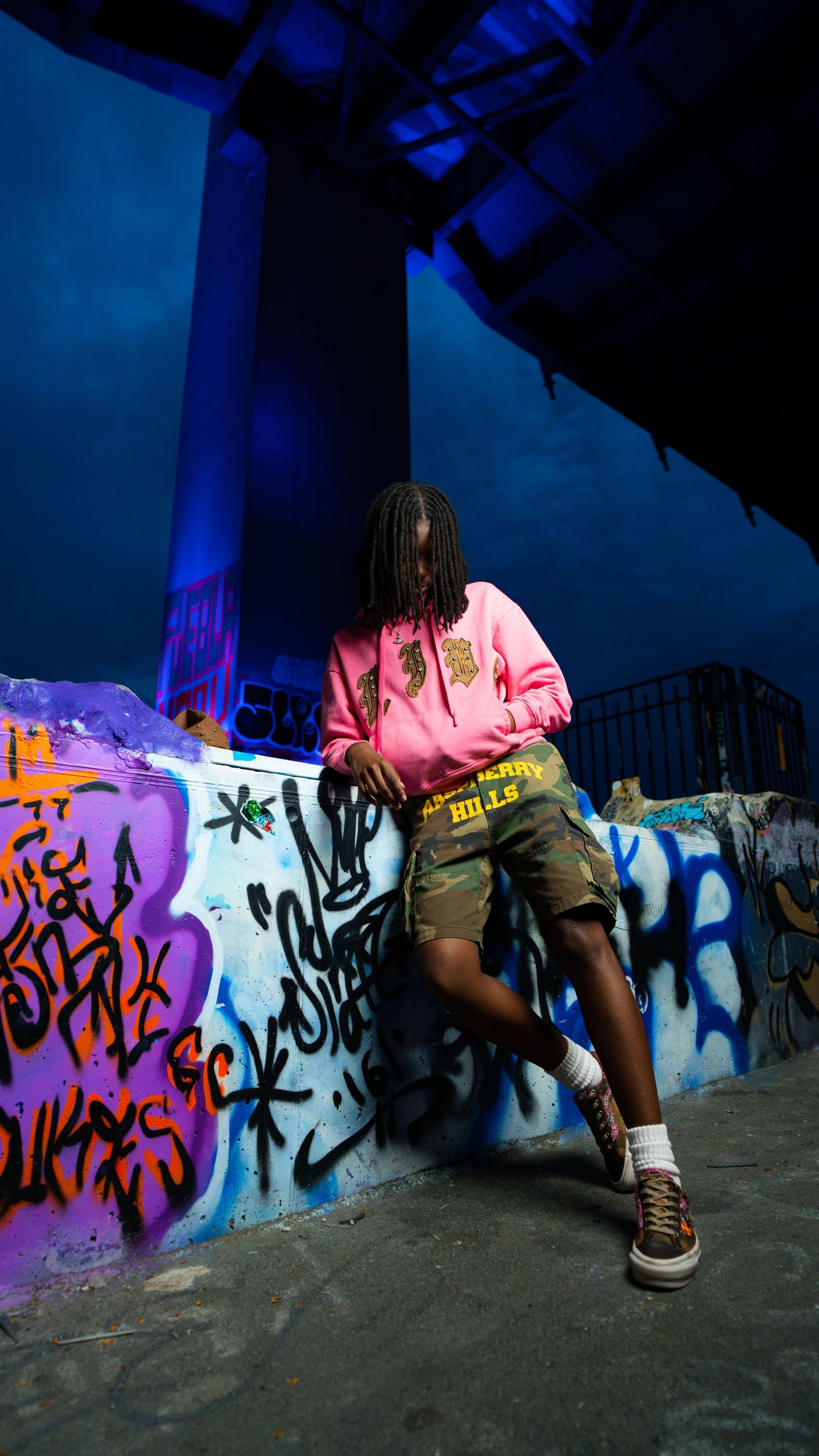 Person in pink hoodie and camo shorts leaning against colorful graffiti wall at night.