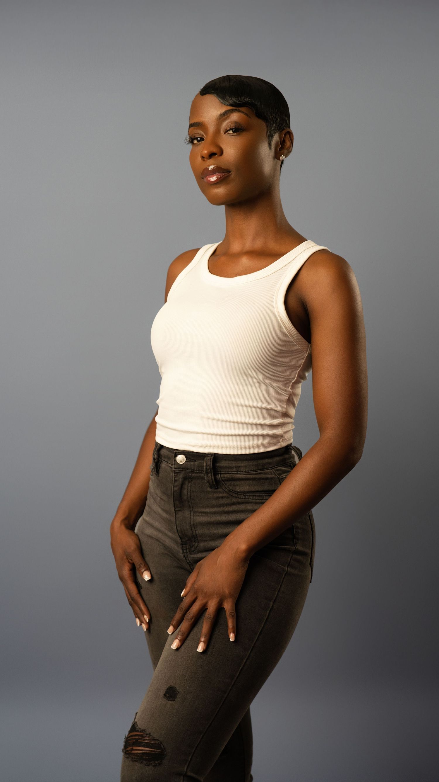 Confident woman in white tank top and dark distressed jeans stands against gray studio background.