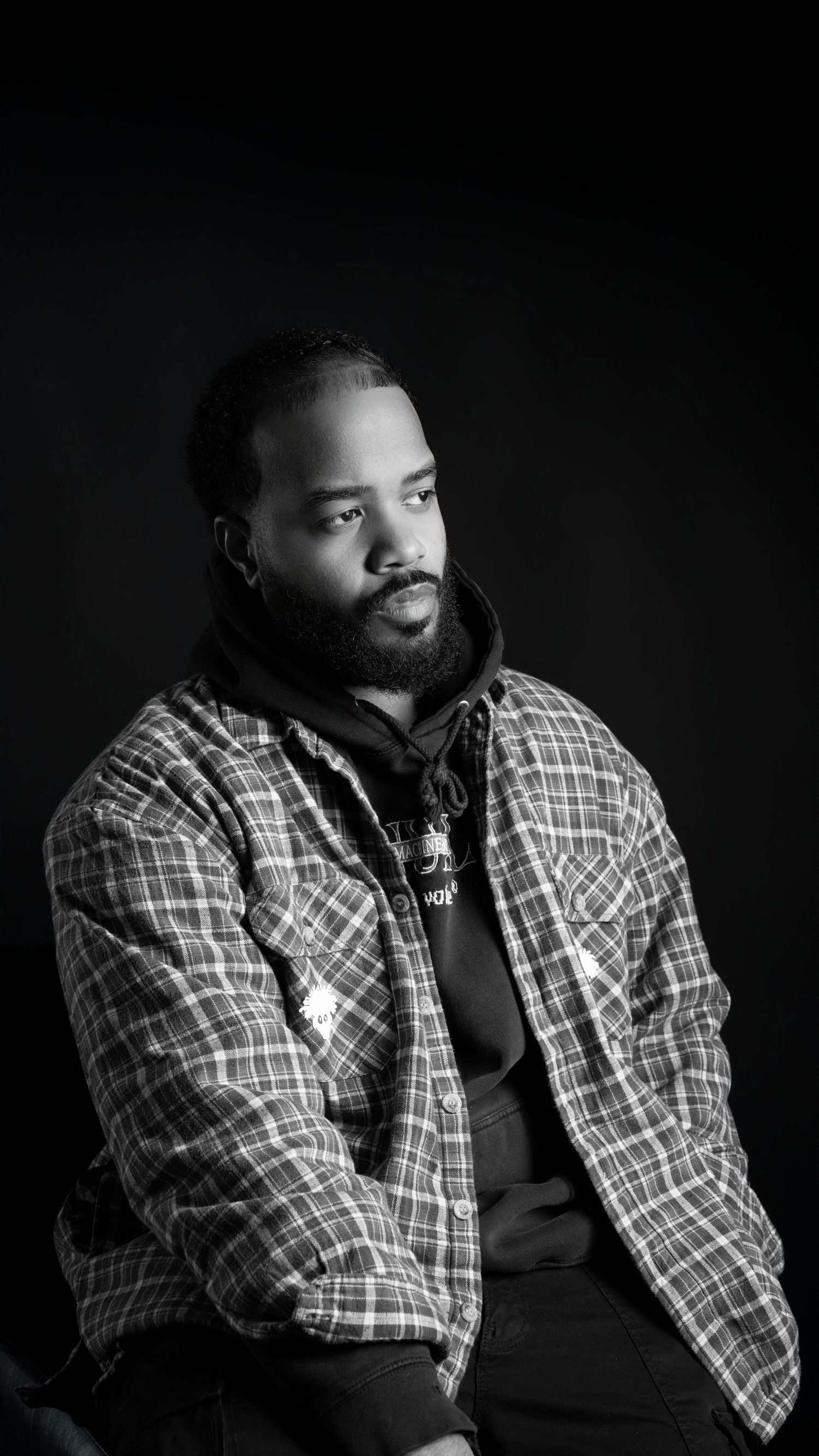 Black and white portrait of bearded man in plaid flannel shirt against dark background.
