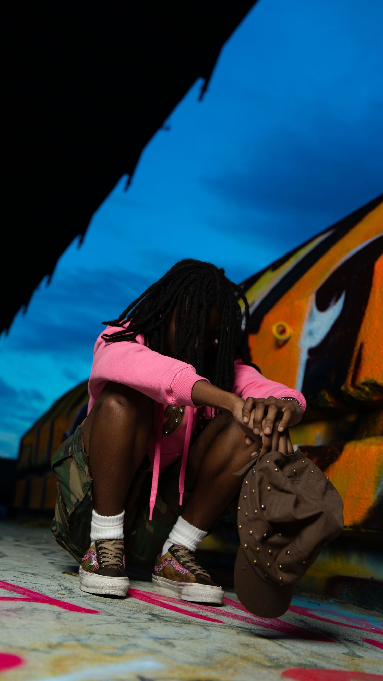 Person in pink hoodie crouches against vibrant graffiti wall holding a studded bucket hat outdoors.