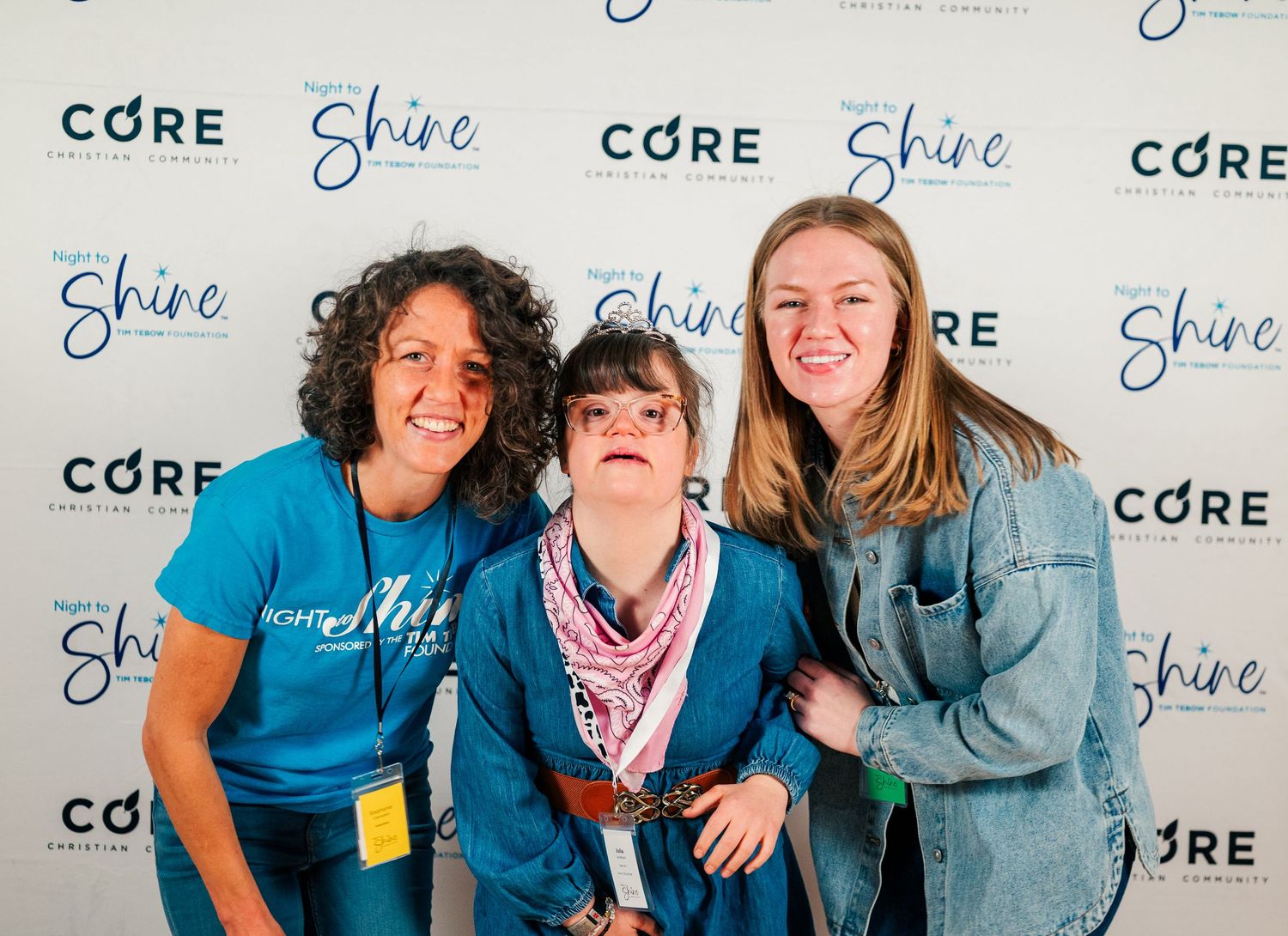 Guest with Down syndrome poses with two volunteers at Night to Shine, smiling in front of event backdrop, celebrating inclusion, joy, and co