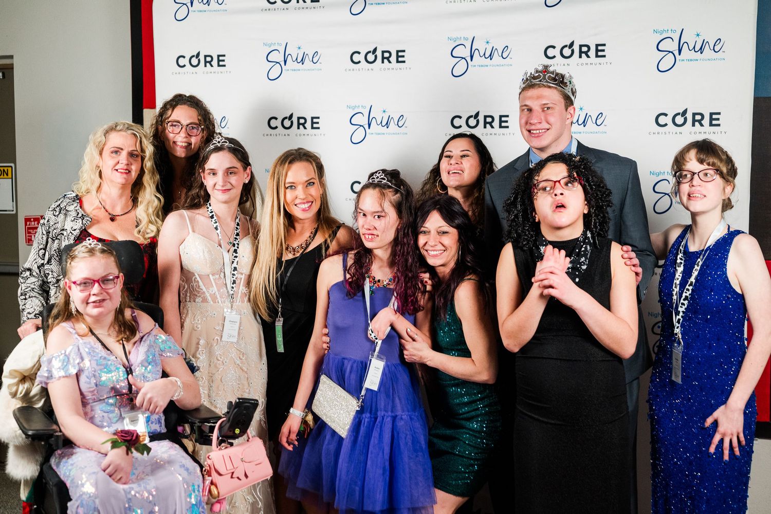 Group portrait at Night to Shine event, guests dressed in formal attire smiling at step-and-repeat backdrop, celebrating an inclusive, joy-f