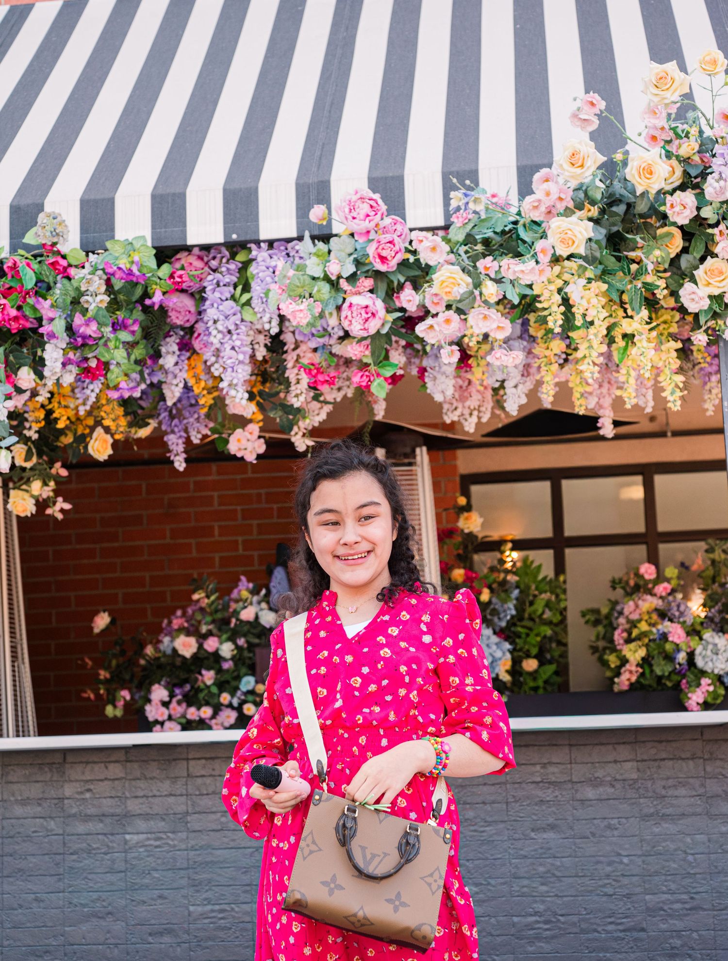 Girl in red dress smiles beneath floral storefront, holding handbag, capturing a bright, stylish moment in a colorful, garden-inspired setti