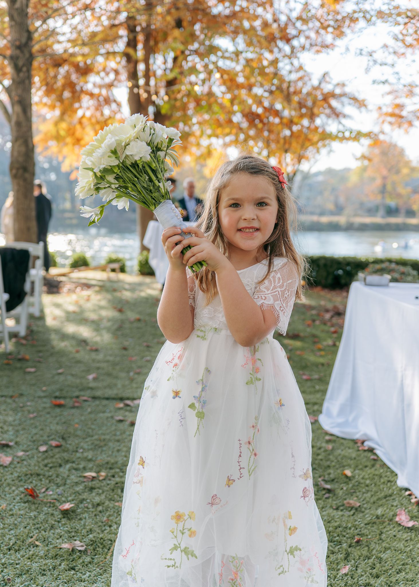 Young flower girl in white floral dress plays with bouquet during autumn wedding.