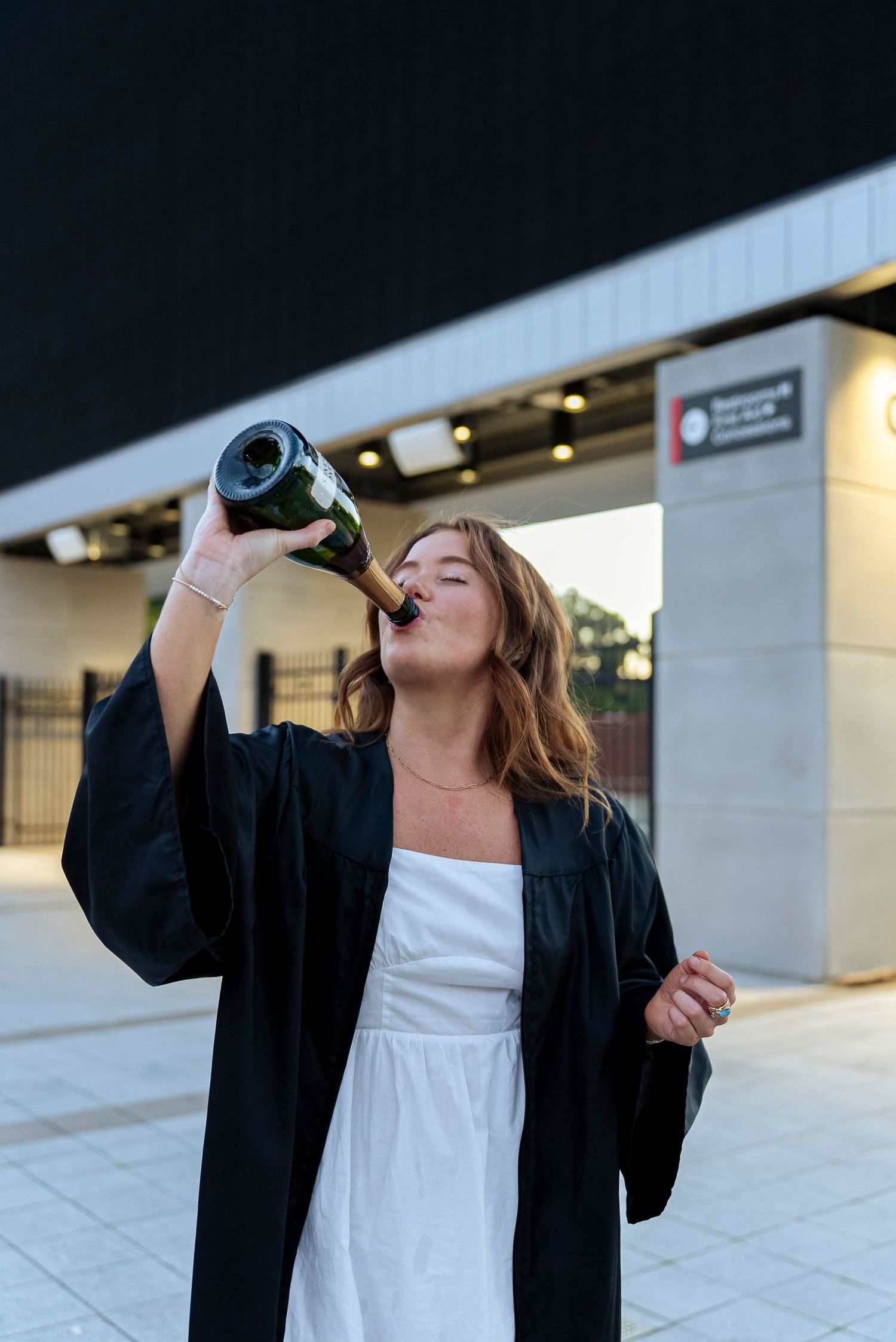 Person in graduation gown takes celebratory drinks outdoors.