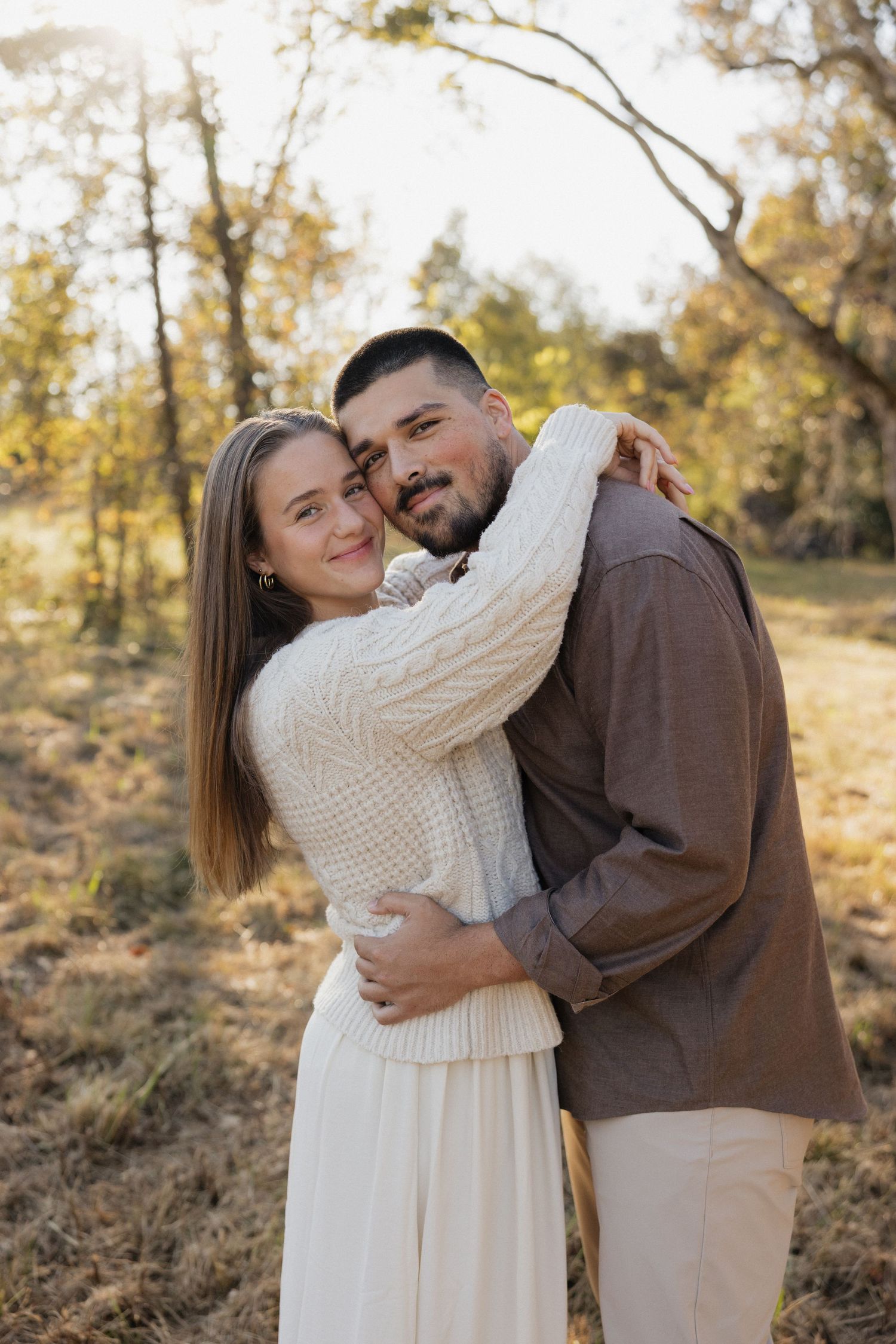 A couple embracing outdoors in warm autumn light wearing neutral colored clothing.