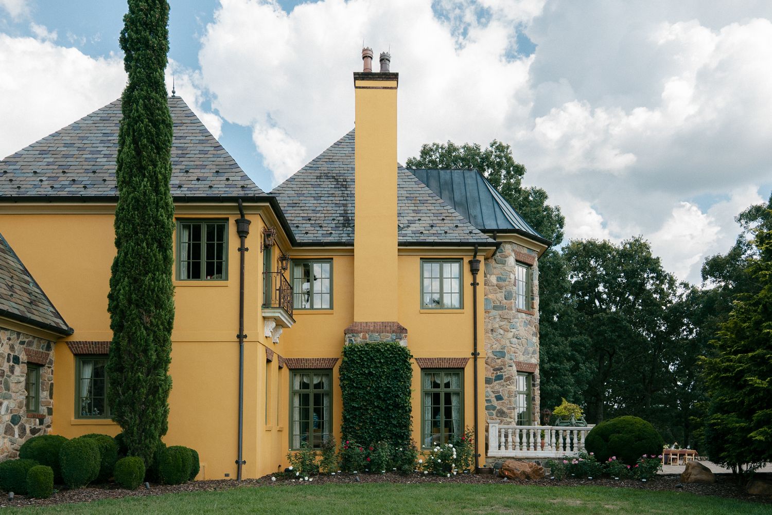 Elegant yellow mansion with stone accents and cypress trees against a cloudy sky.
