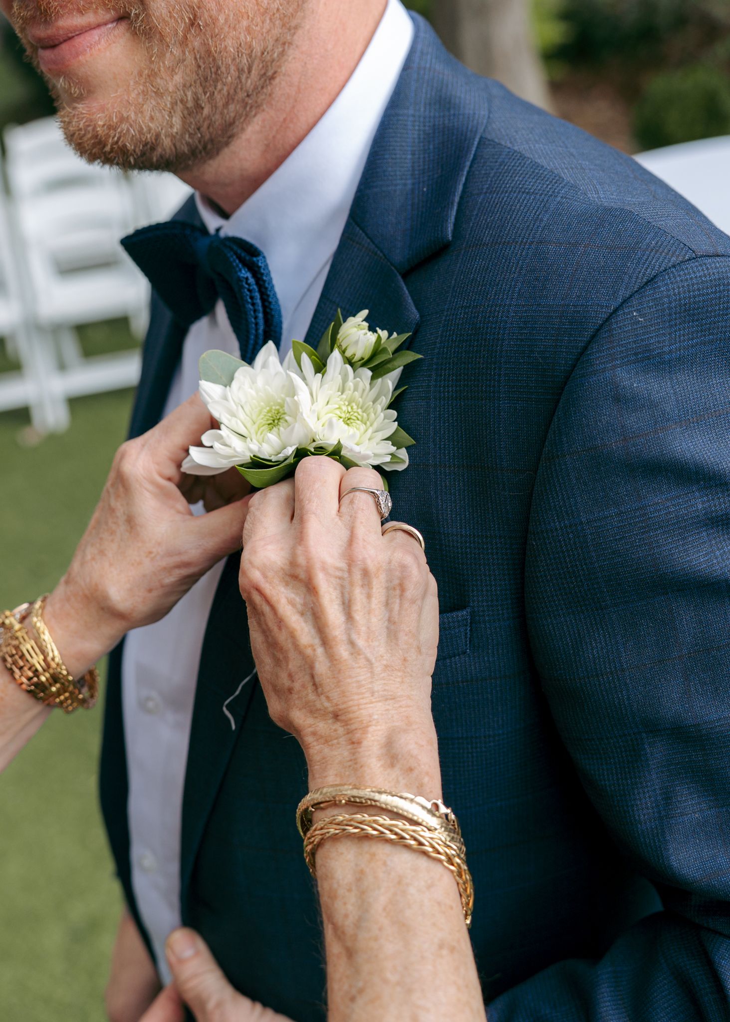 Close-up of boutonniere being pinned to navy suit jacket with gold bracelet visible.