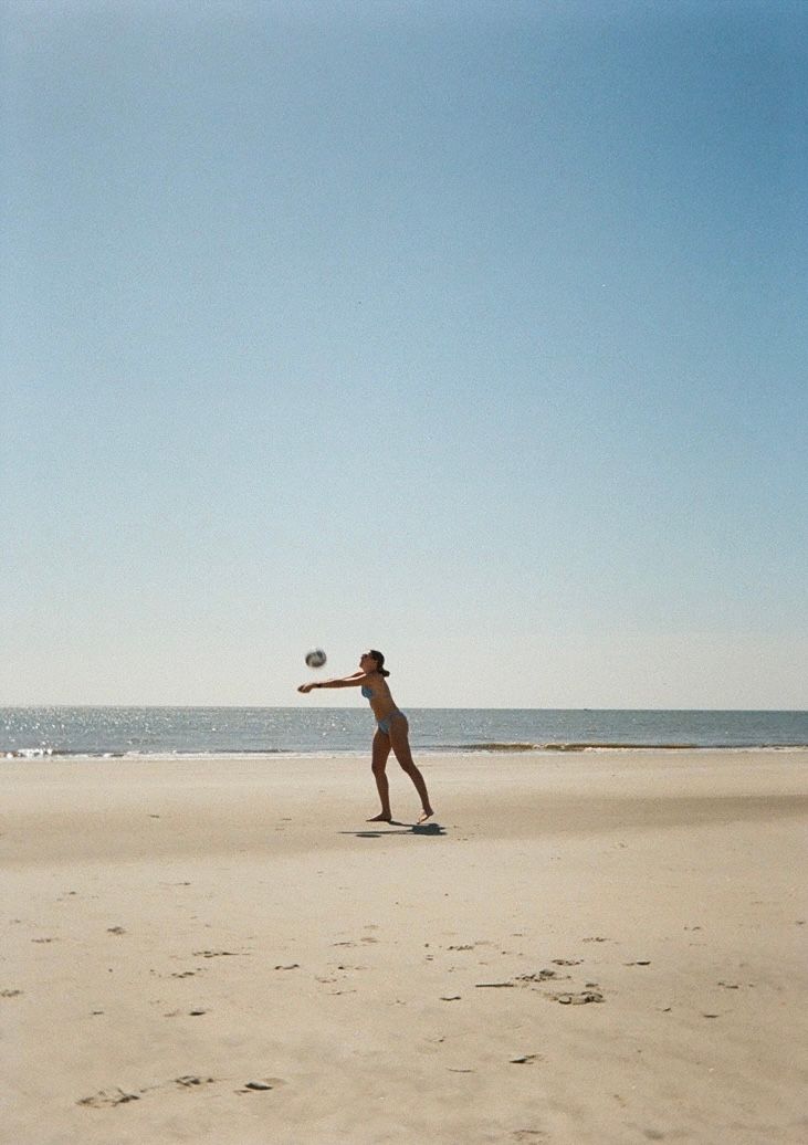 Silhouettes of people playing with a ball on an expansive empty beach against a blue sky.