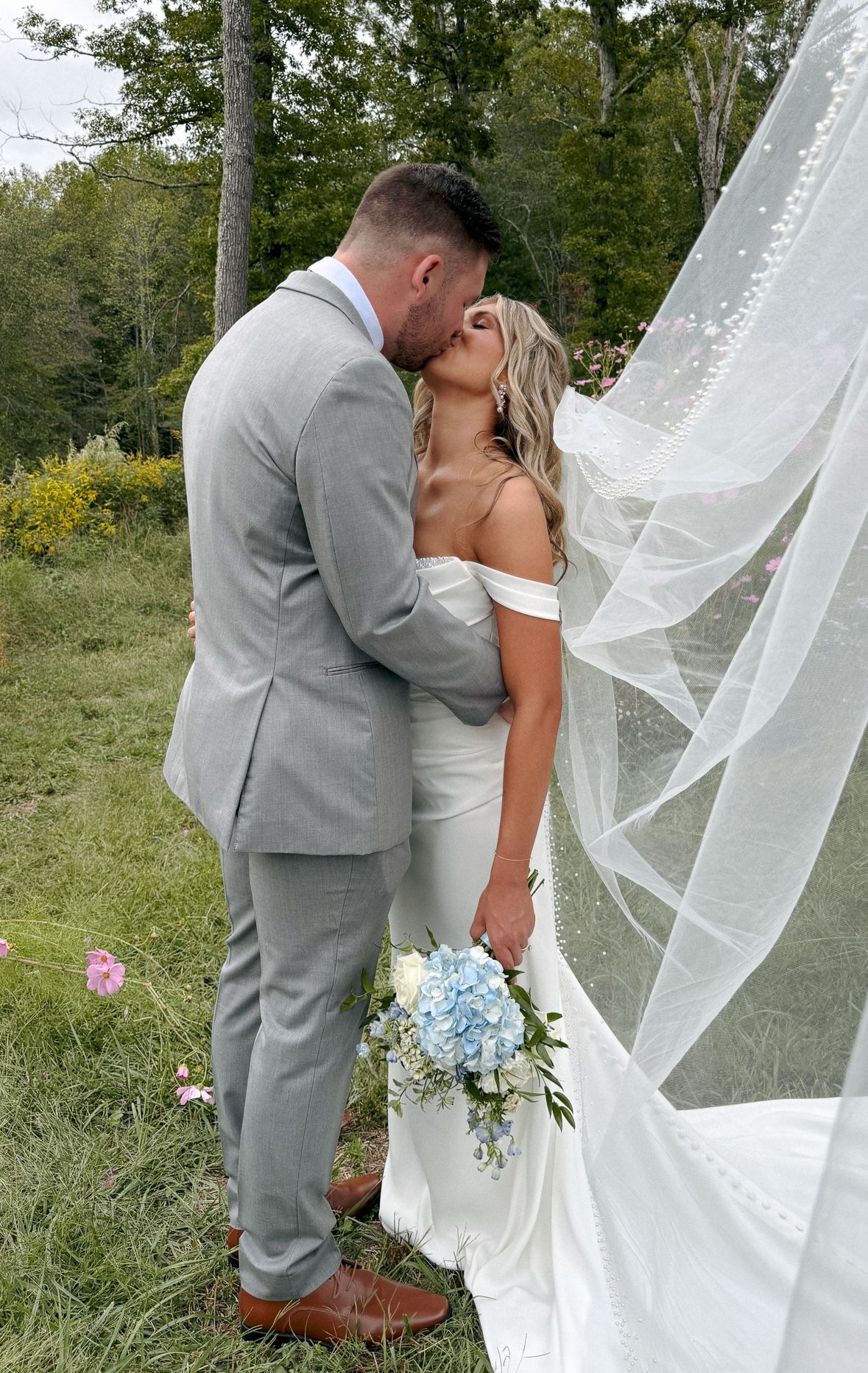 A romantic wedding moment captured outdoors with a flowing veil and blue bouquet.