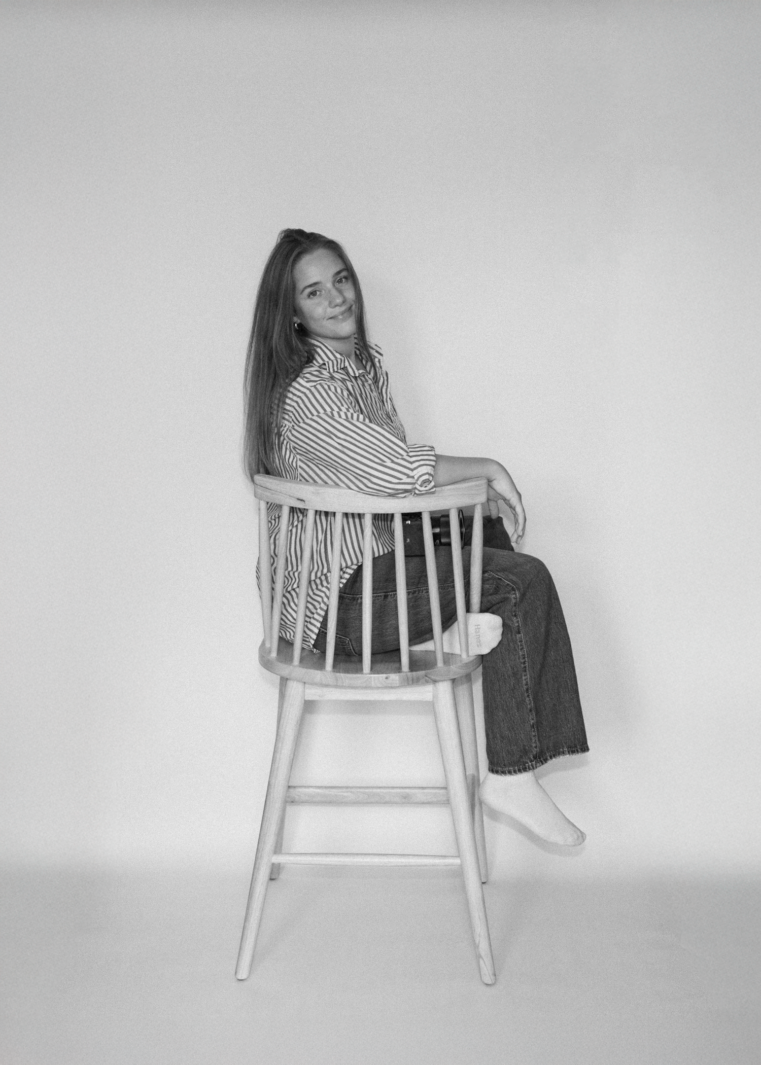 Black and white portrait of someone posed on a wooden chair against a plain background.