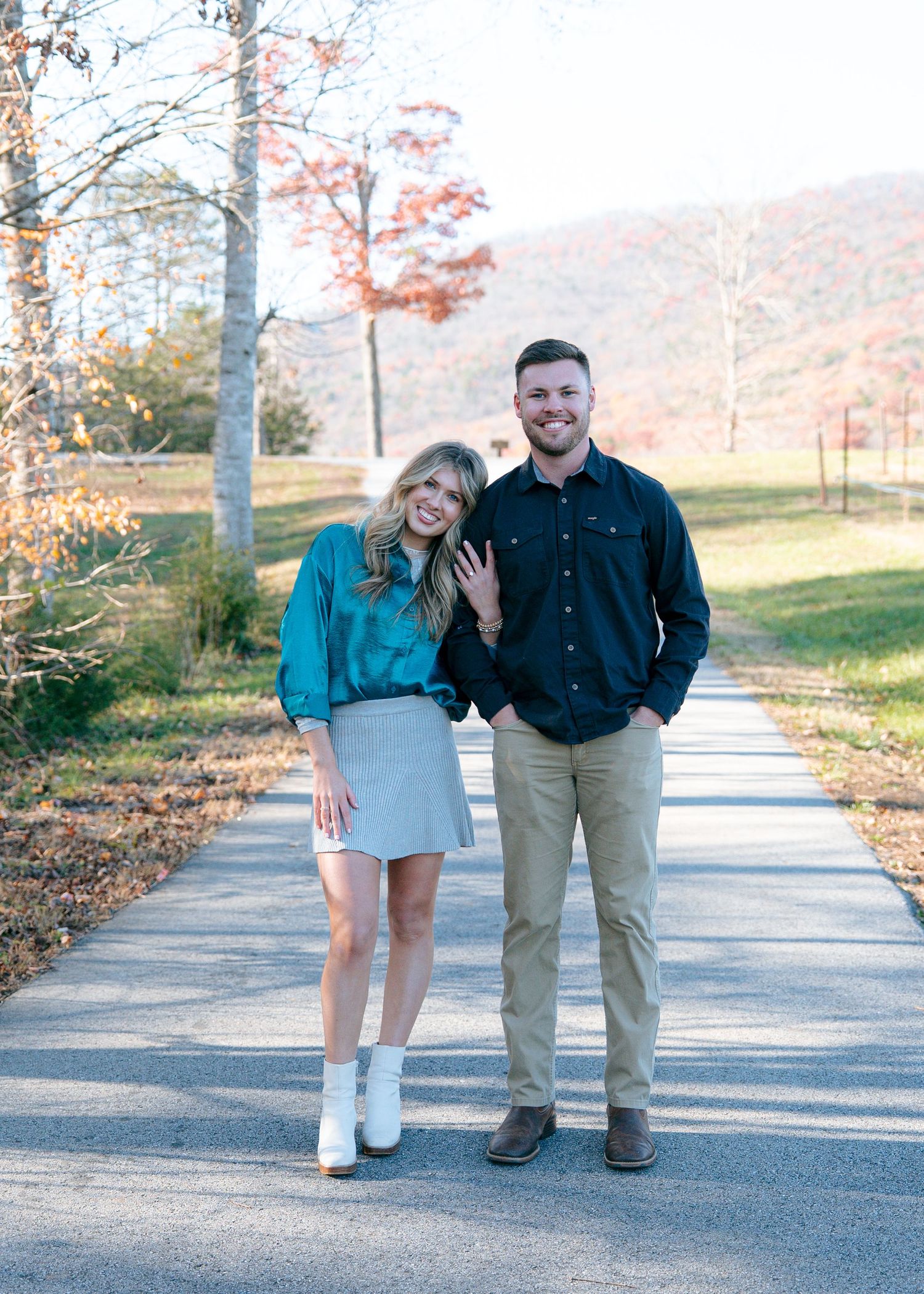 Couple posing playfully on a paved path during autumn with colorful foliage in background.