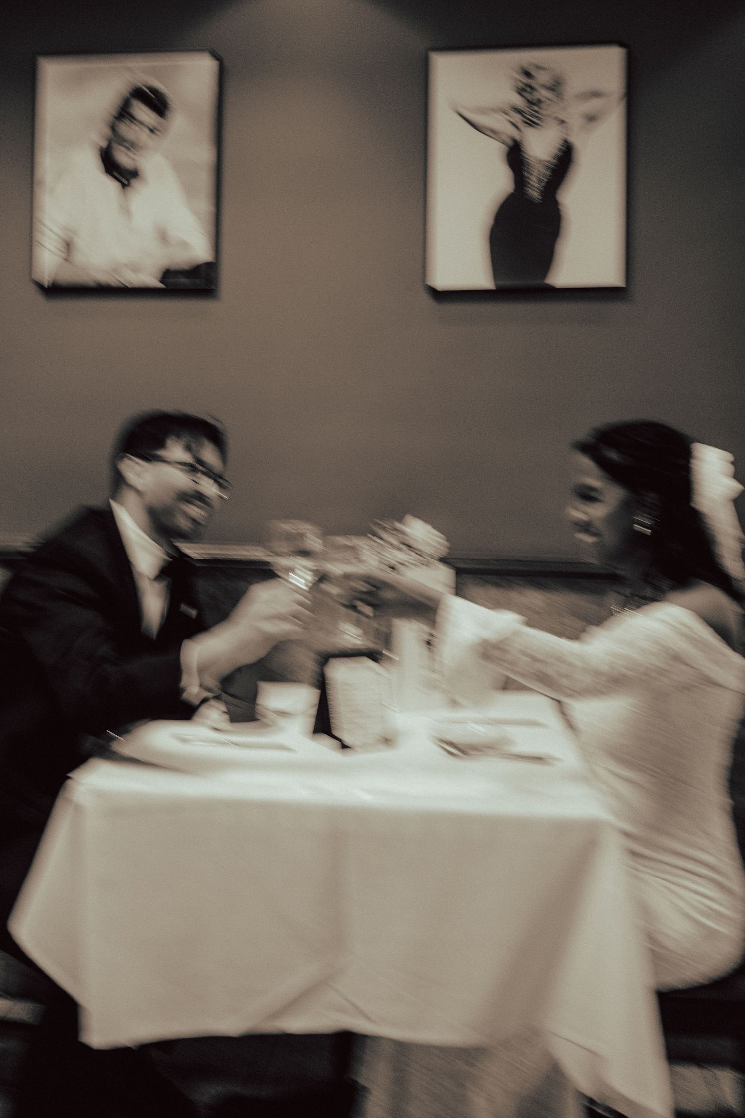 Blurred vintage-style photo of a couple dining at a table with white tablecloth and artwork on dark walls.