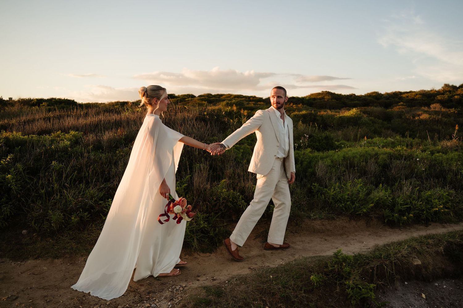 Bride in a flowing white gown and veil holding a bouquet walks hand in hand with a groom in a cream suit along a coastal path at sunset.