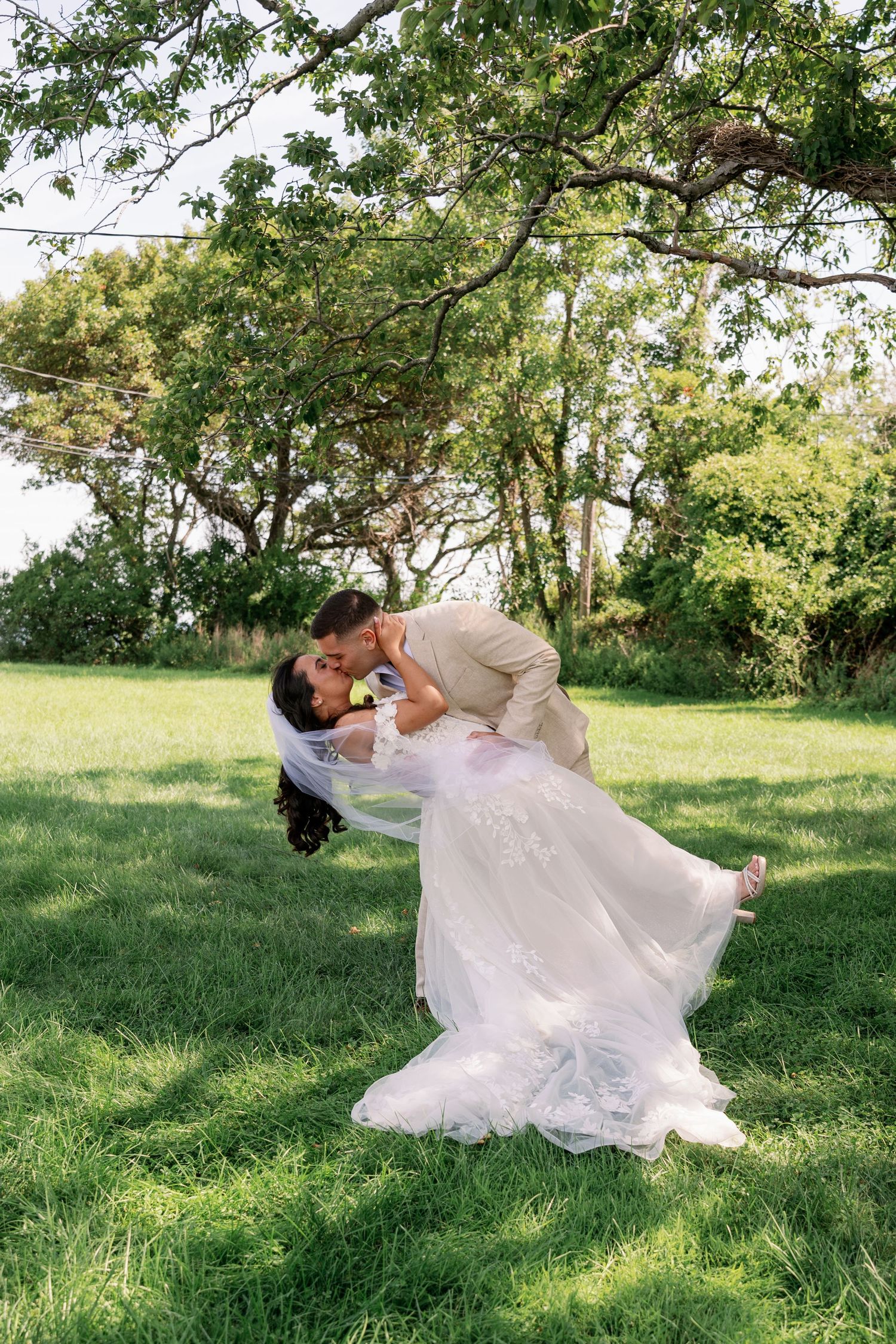 Groom in a tan suit dips and kisses his bride in a flowing lace wedding gown and veil on a sunny green lawn beneath tall trees.