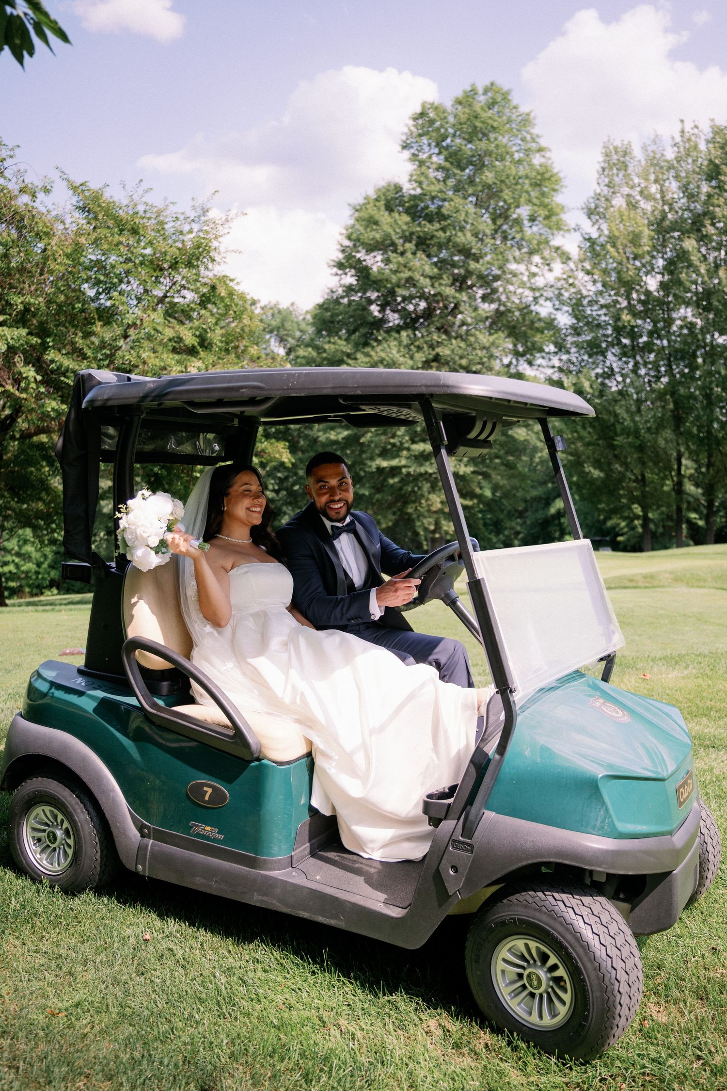 Bride and groom riding a green golf cart on a sunny day, bride holding a white bouquet in a lush garden setting.