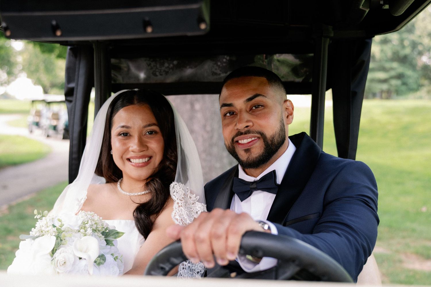 Smiling bride and groom riding a golf cart on their wedding day, bride in white gown with bouquet, groom in navy suit.