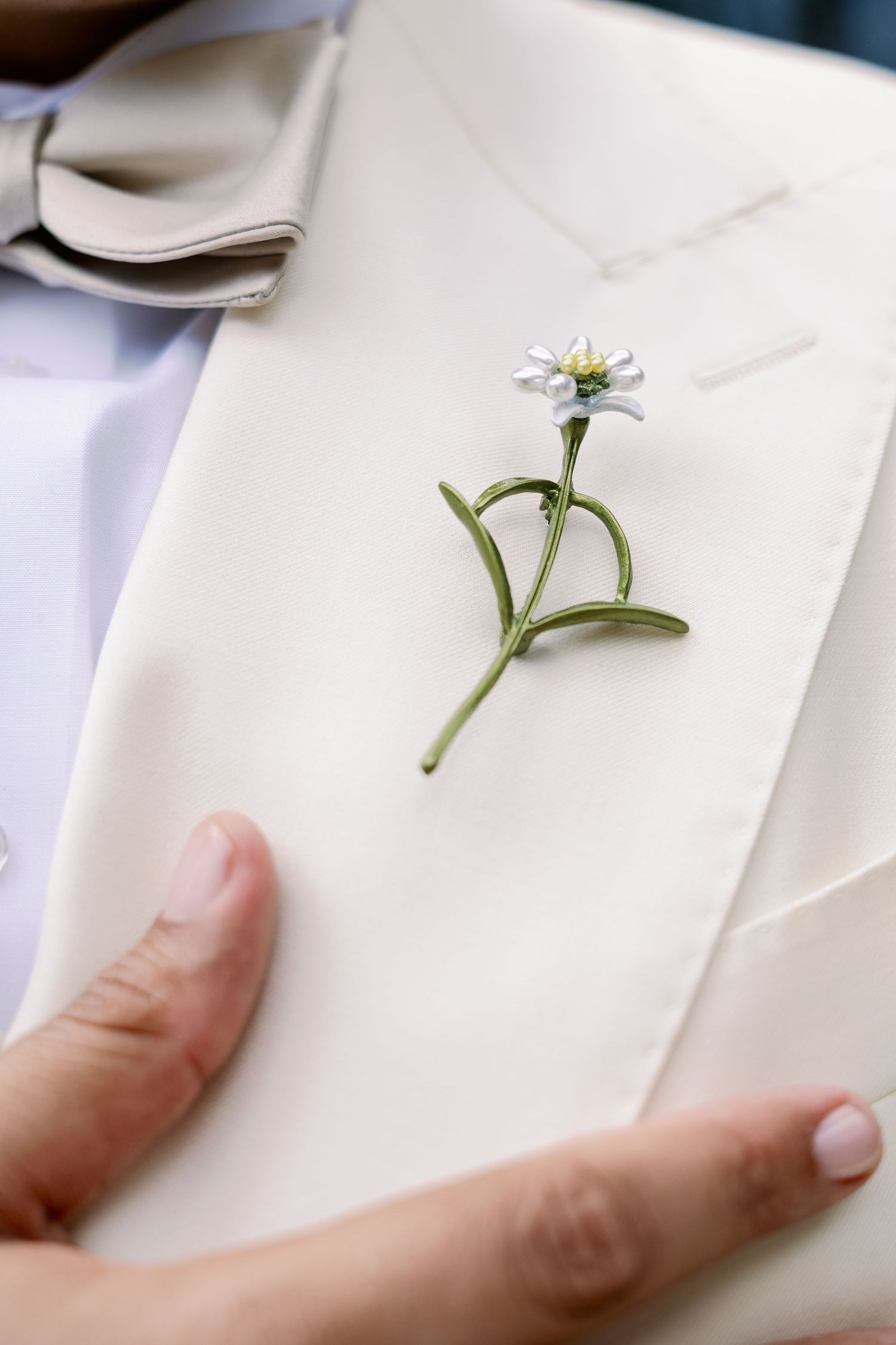 Close-up of a daisy boutonniere on a cream wedding suit lapel, with a hand gently adjusting it.