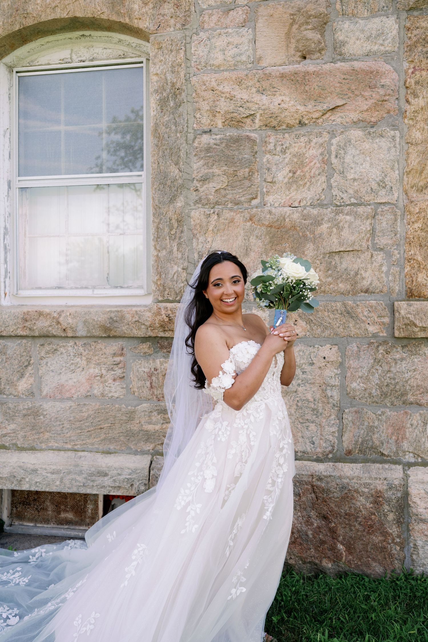 Smiling bride in an off-shoulder lace wedding gown and veil holds a white floral bouquet beside a rustic stone building.