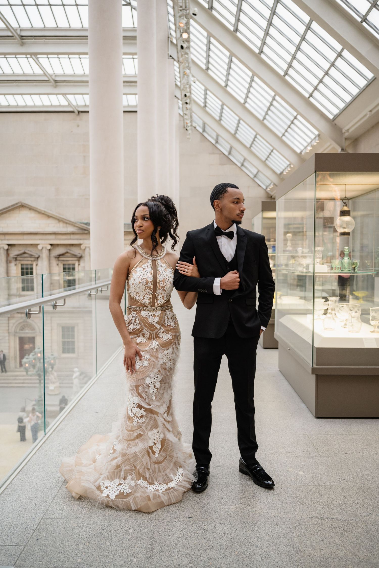 A couple in formal wedding attire poses playfully in a bright museum gallery with skylights and white walls.