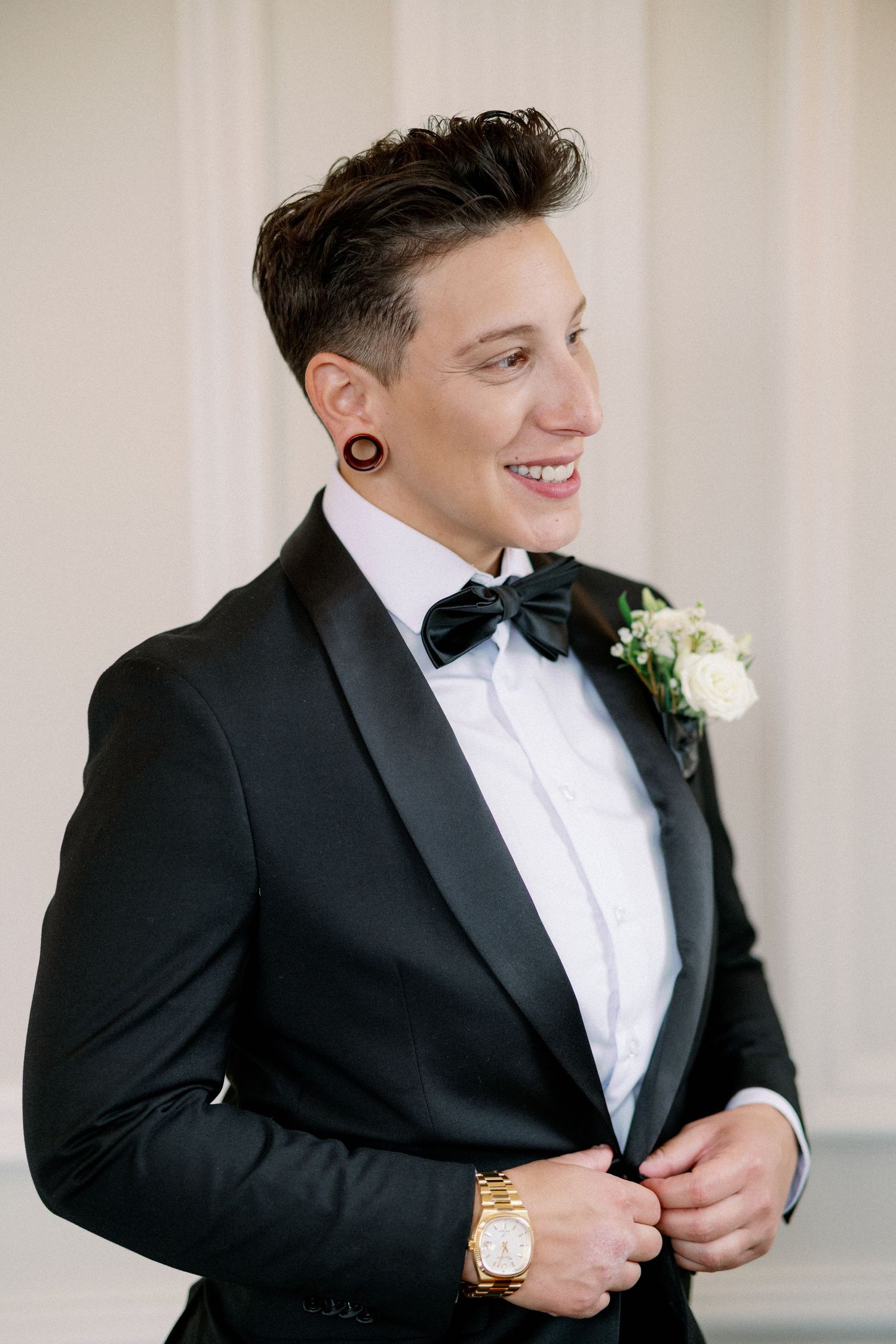 Smiling groom in black tuxedo with bow tie and white boutonniere, adjusting jacket, wearing a gold watch.