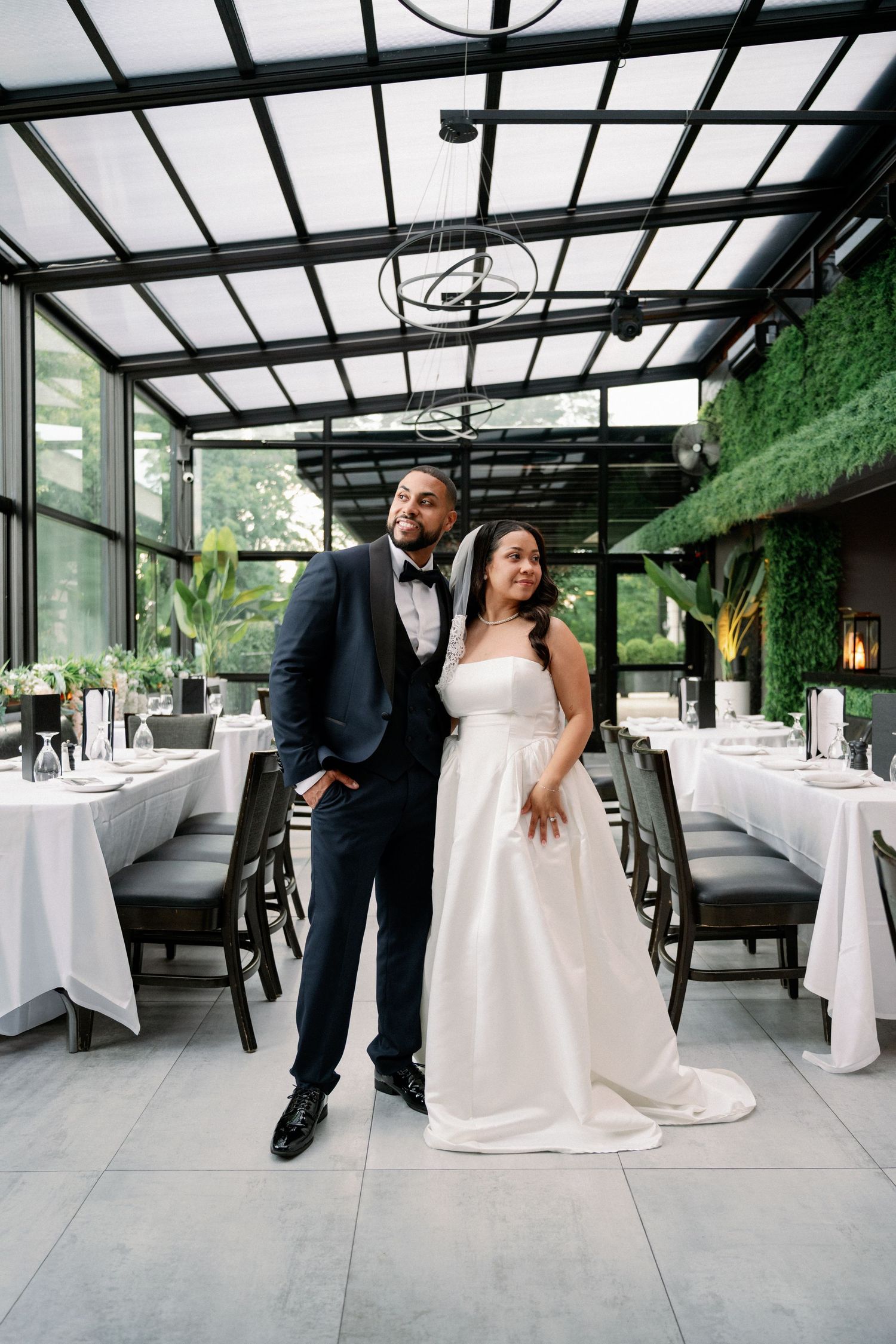 Bride and groom pose elegantly in a glass-ceiling wedding venue with lush greenery walls and white-draped reception tables.