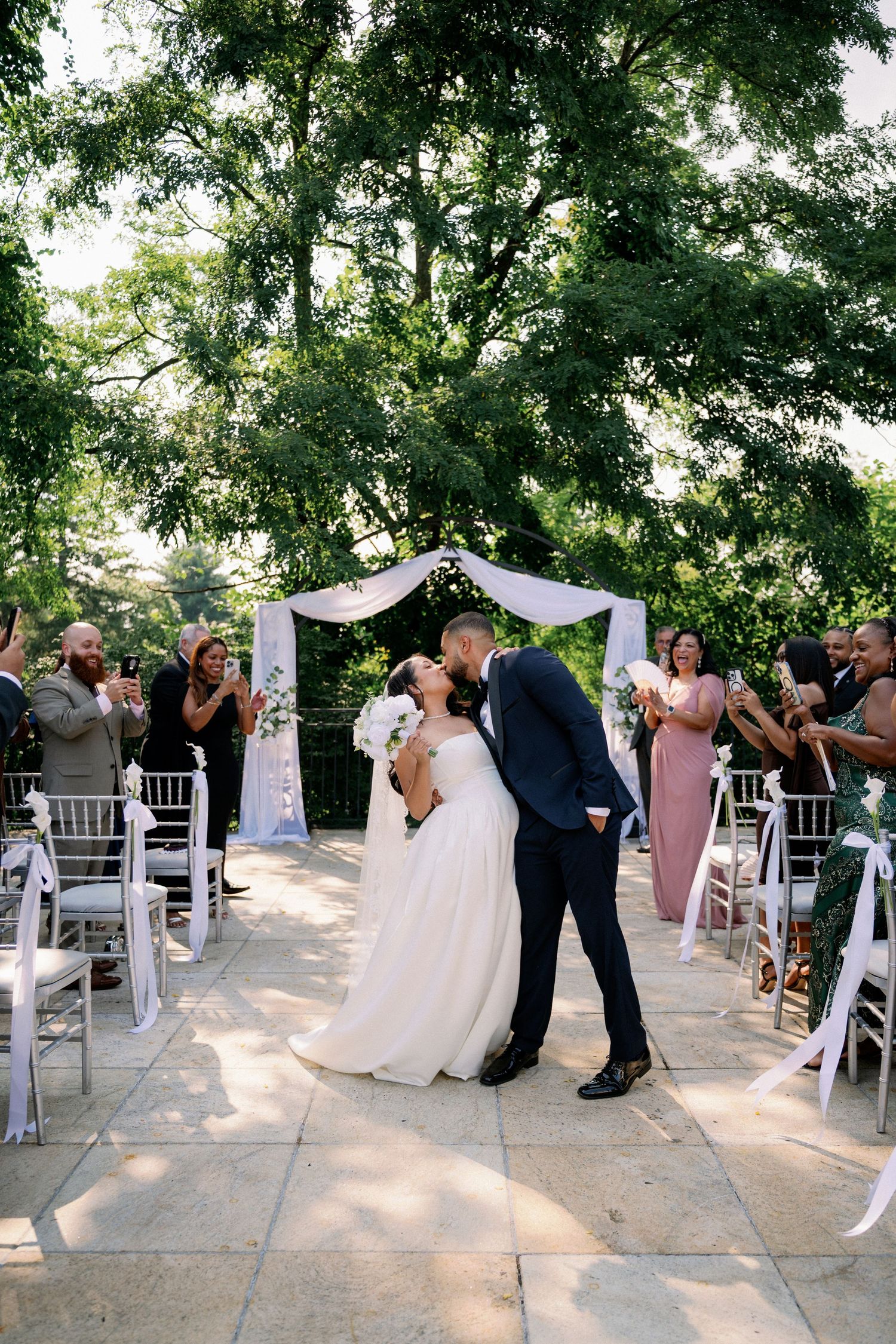 Bride and groom share first kiss at outdoor wedding ceremony with white arch and cheering guests.