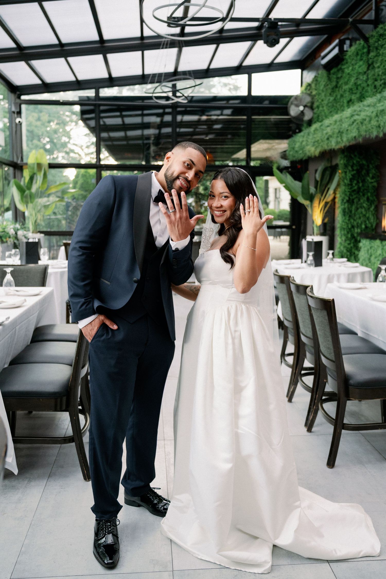 Bride and groom showing wedding rings, smiling in elegant venue with greenery walls and white tablecloths.