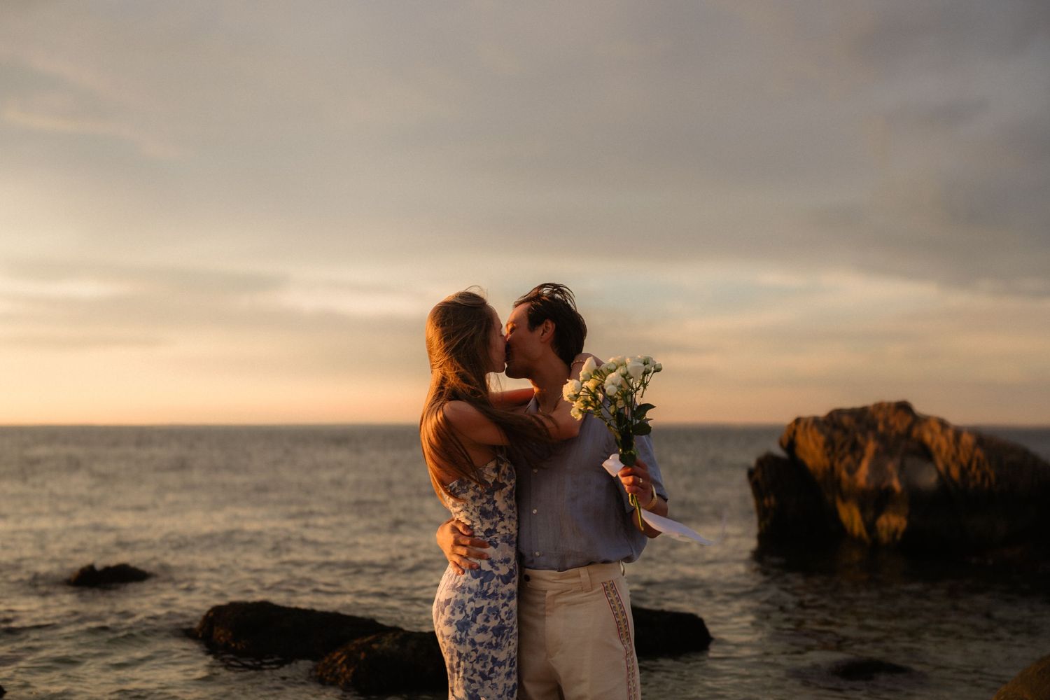 Silhouetted couple embraces on beach during golden sunset with dramatic clouds and ocean waves in background.