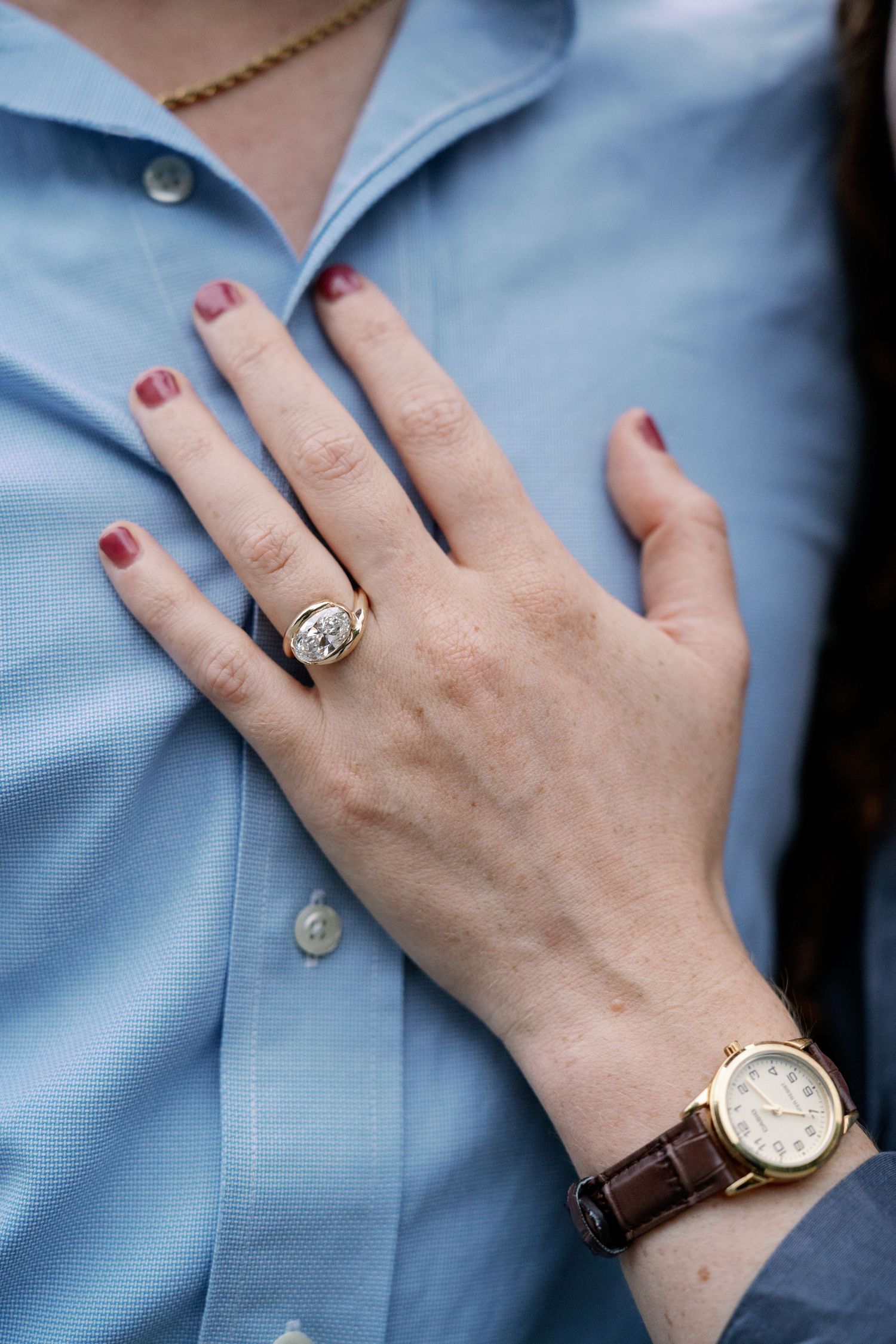 Woman's hand with red manicure and a diamond engagement ring resting on a man's blue button-up shirt, with a gold wristwatch on her wrist.