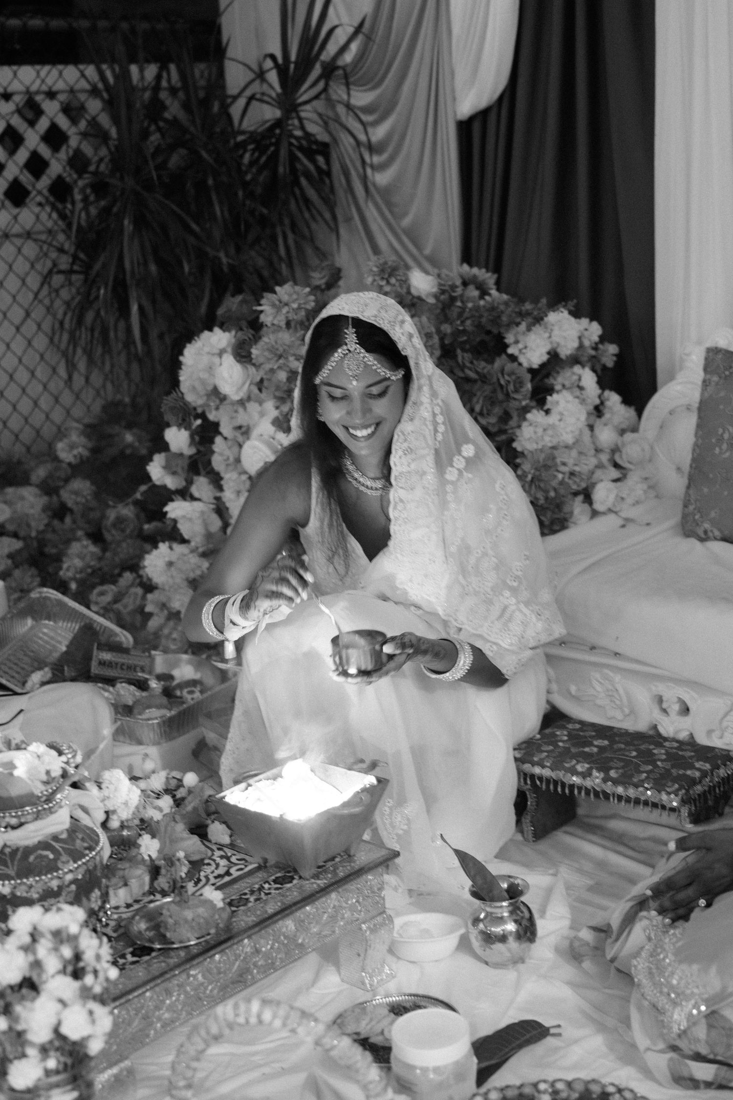 Smiling bride in a white lace veil and headpiece kneels beside a glowing ceremonial fire at a traditional wedding, surrounded by white flowe