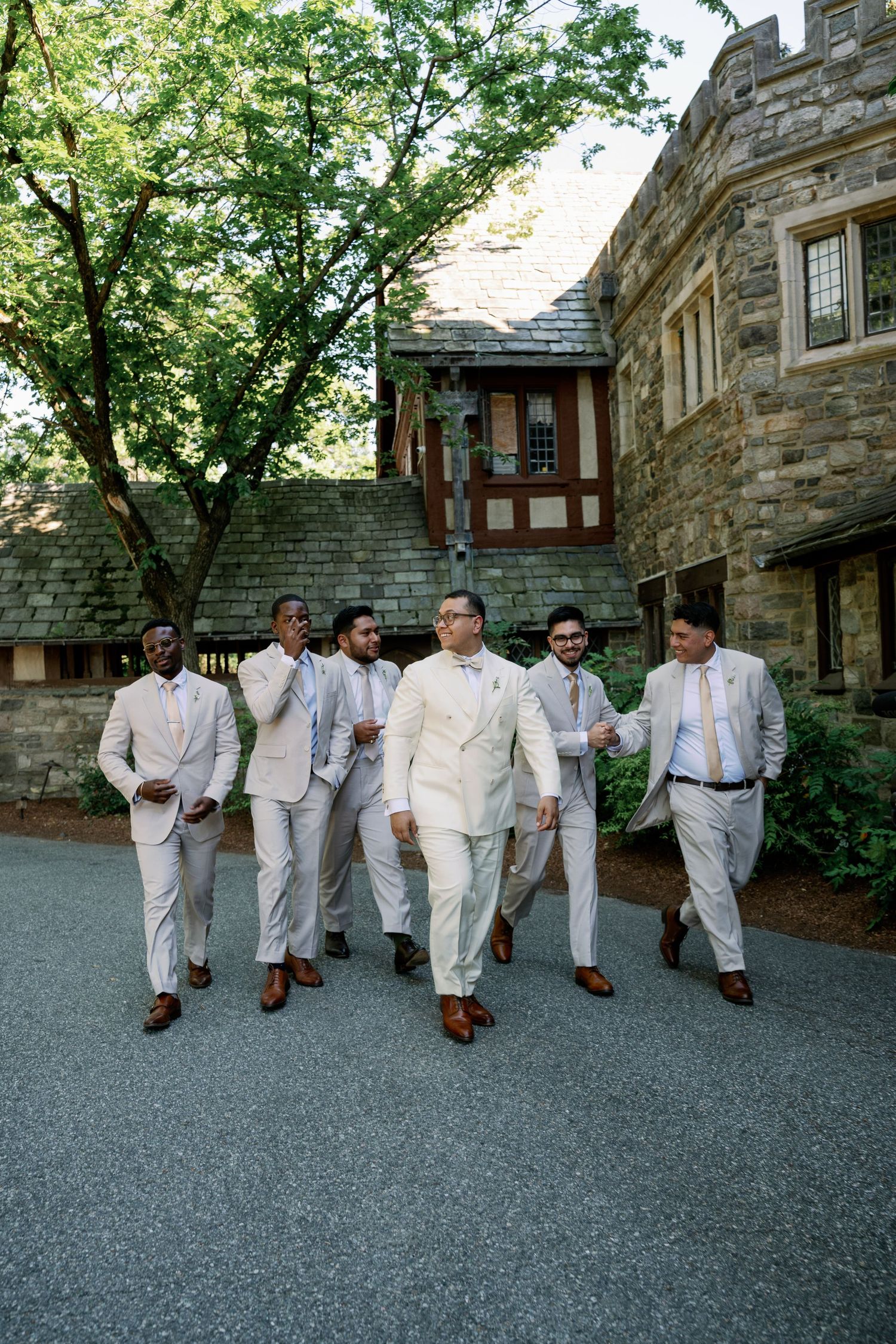Groom and groomsmen in cream suits with brown shoes walk outside a stone mansion on a wedding day.