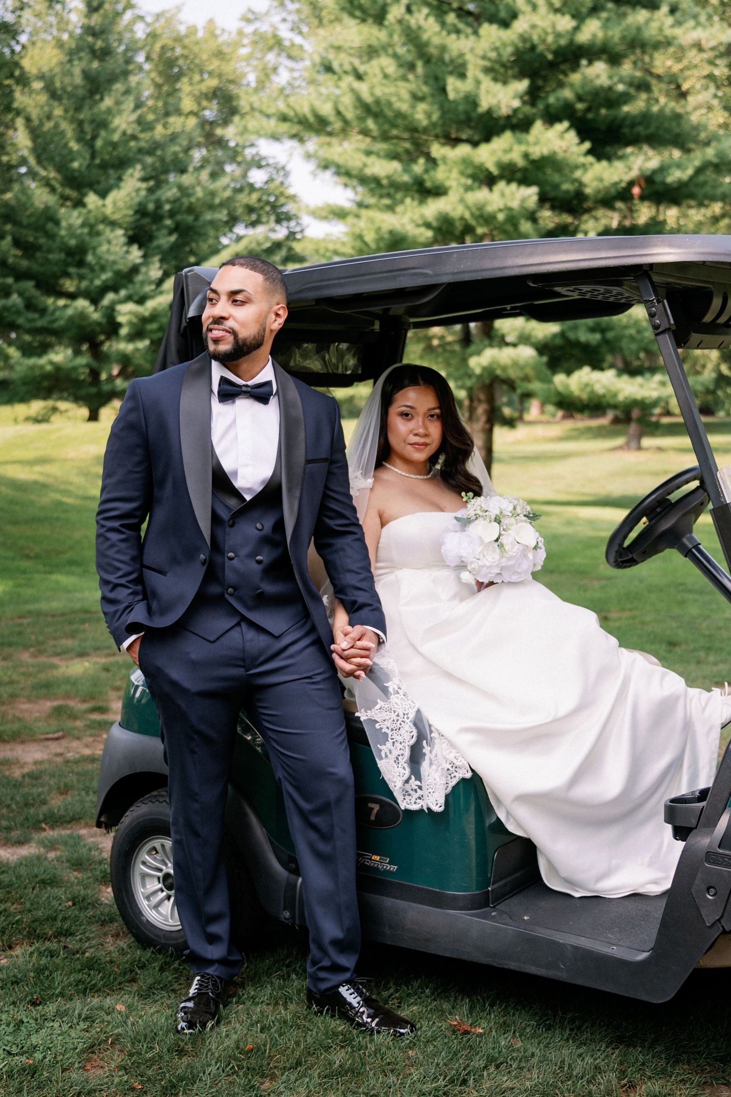Bride and groom pose by a golf cart on lush green grounds, she in a white gown, he in a navy tuxedo.