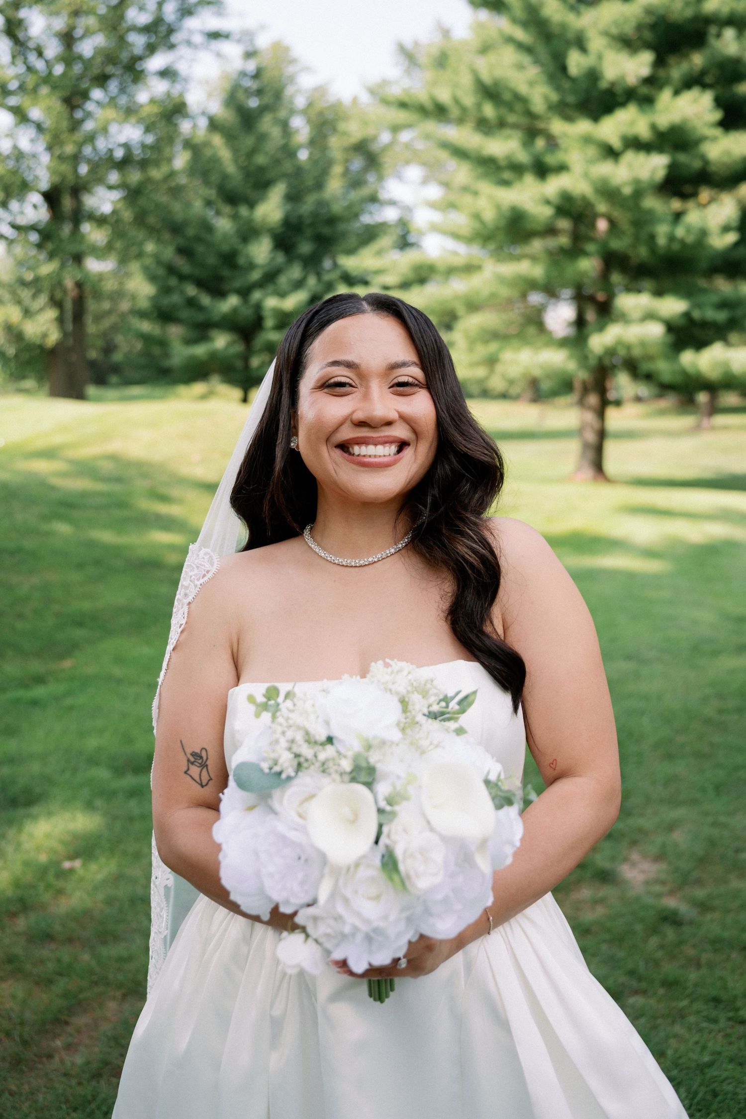 Smiling bride in strapless white gown holds white floral bouquet outdoors with lush green trees behind her.