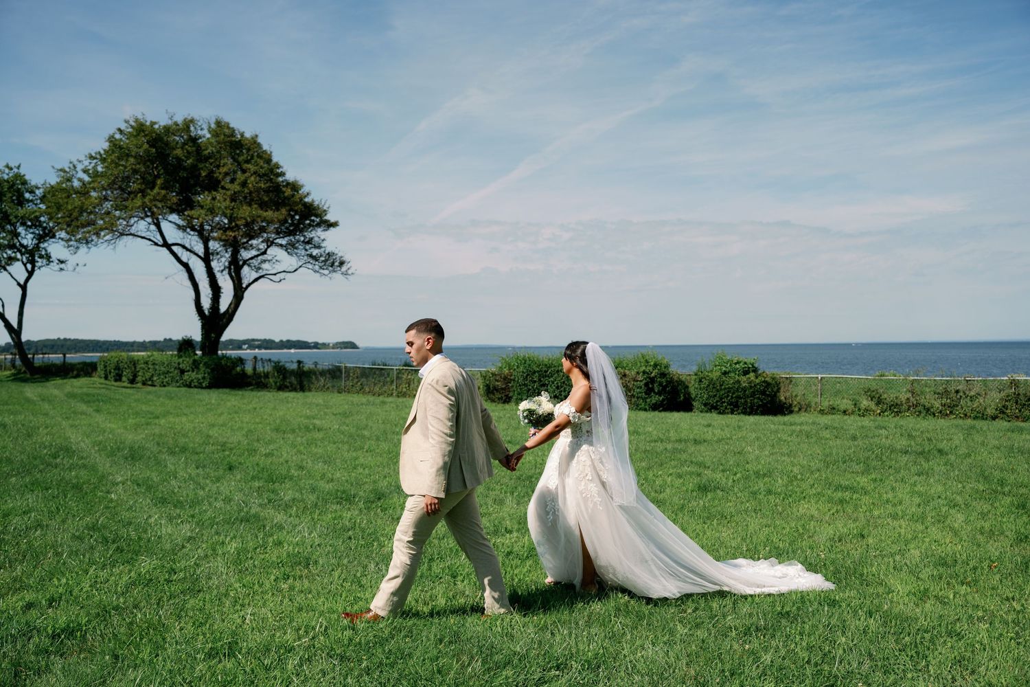 Bride in a long white veil and gown holds hands with a groom in a light suit while walking across a green lawn near the waterfront.
