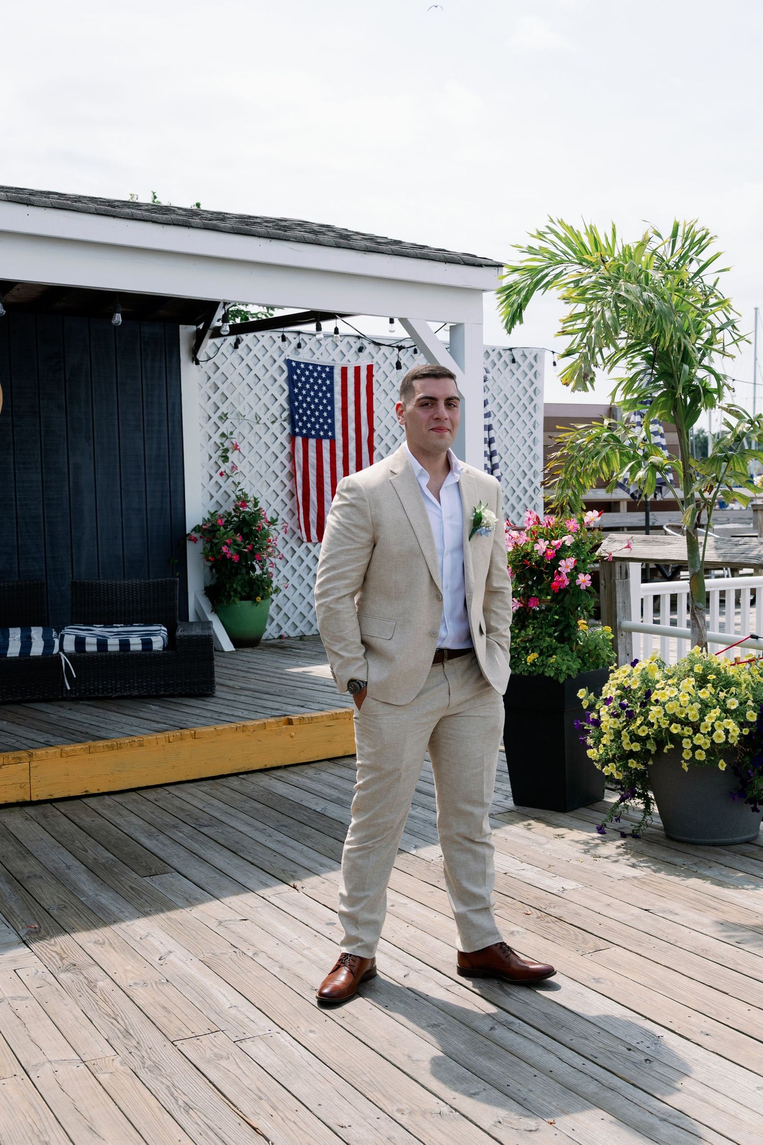 Groom in a tan suit with a boutonniere stands on a wooden deck near an American flag, flowering planters, and a palm tree.