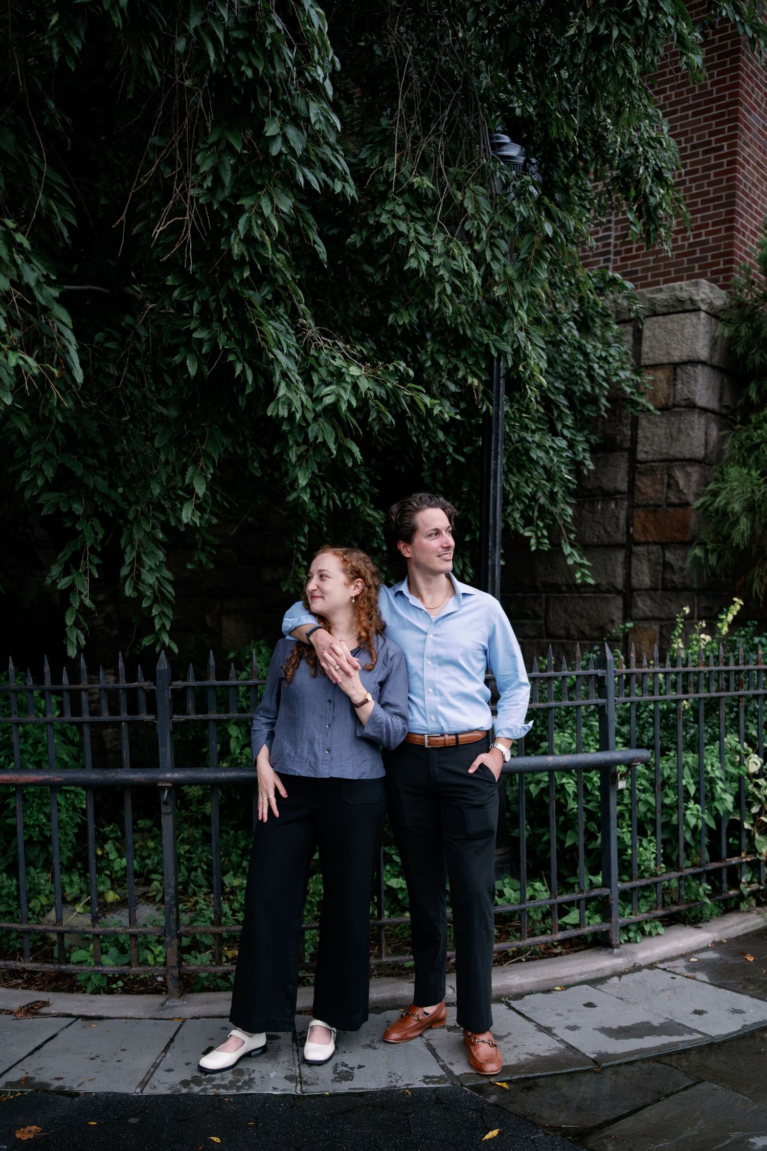 Couple embracing and smiling on a wet sidewalk beside an iron fence, with ivy-covered brick and stone behind them.