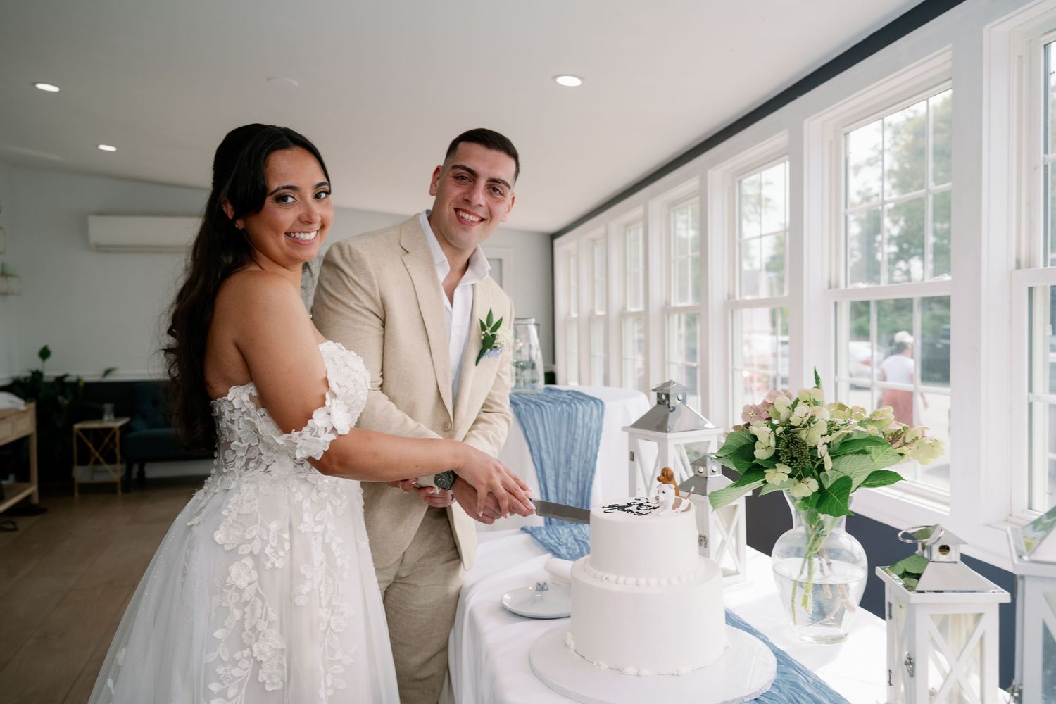 Bride in a lace wedding gown and groom in a tan suit cut their white wedding cake together in a bright room with large windows.