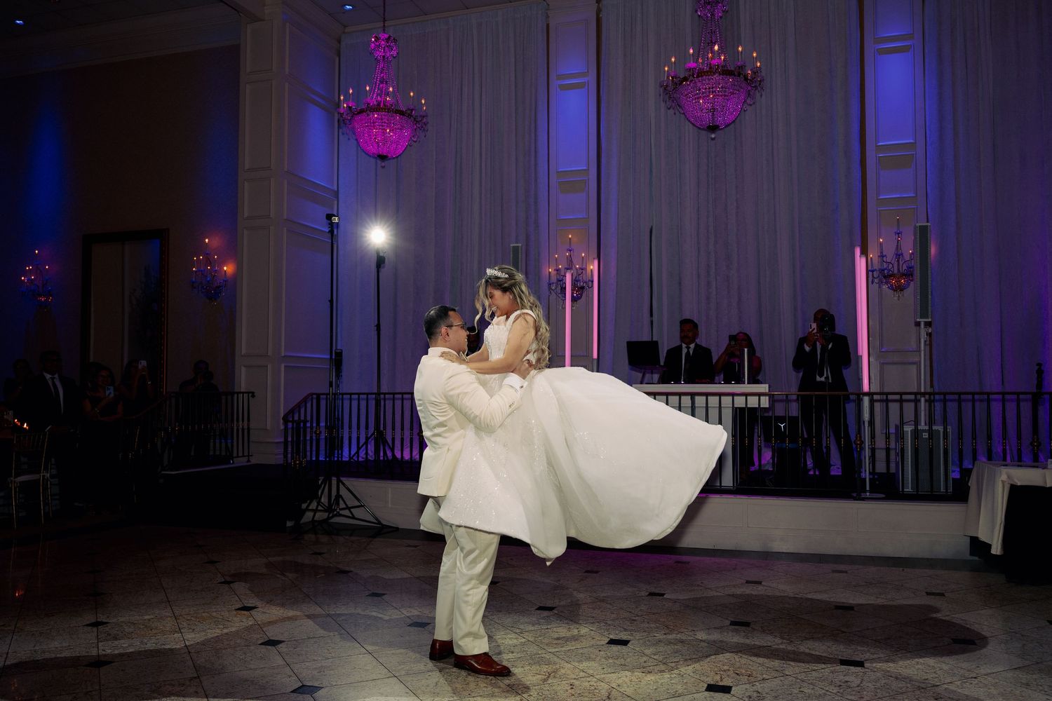 Groom lifts bride in white gown during romantic first dance at elegant wedding reception with purple lighting.
