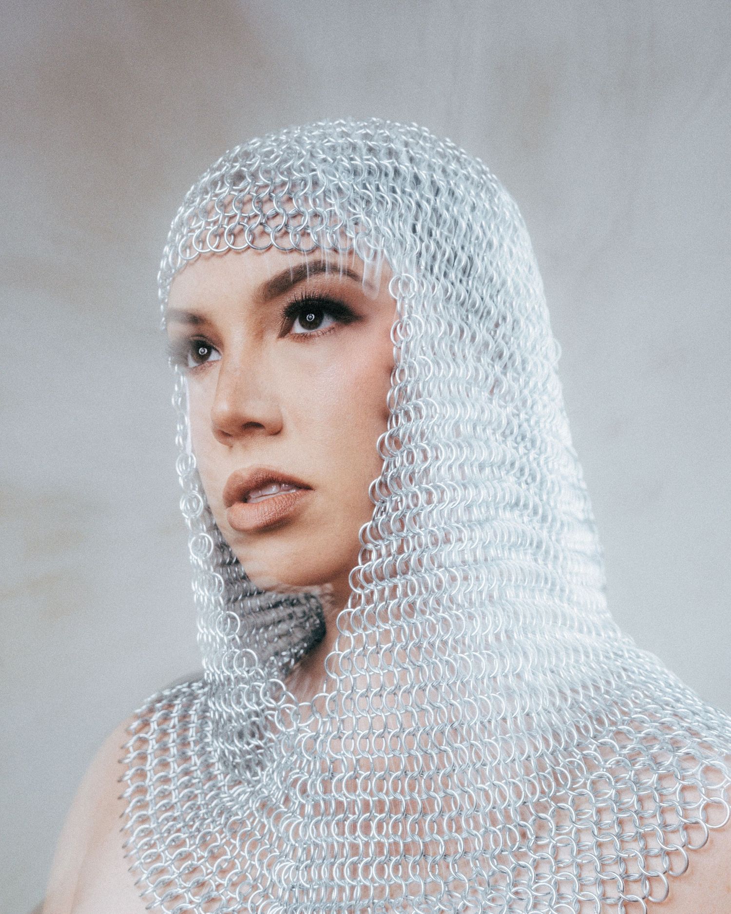 Woman wearing silver chainmail coif hood draping over shoulders, medieval armor headpiece, fashion editorial portrait.