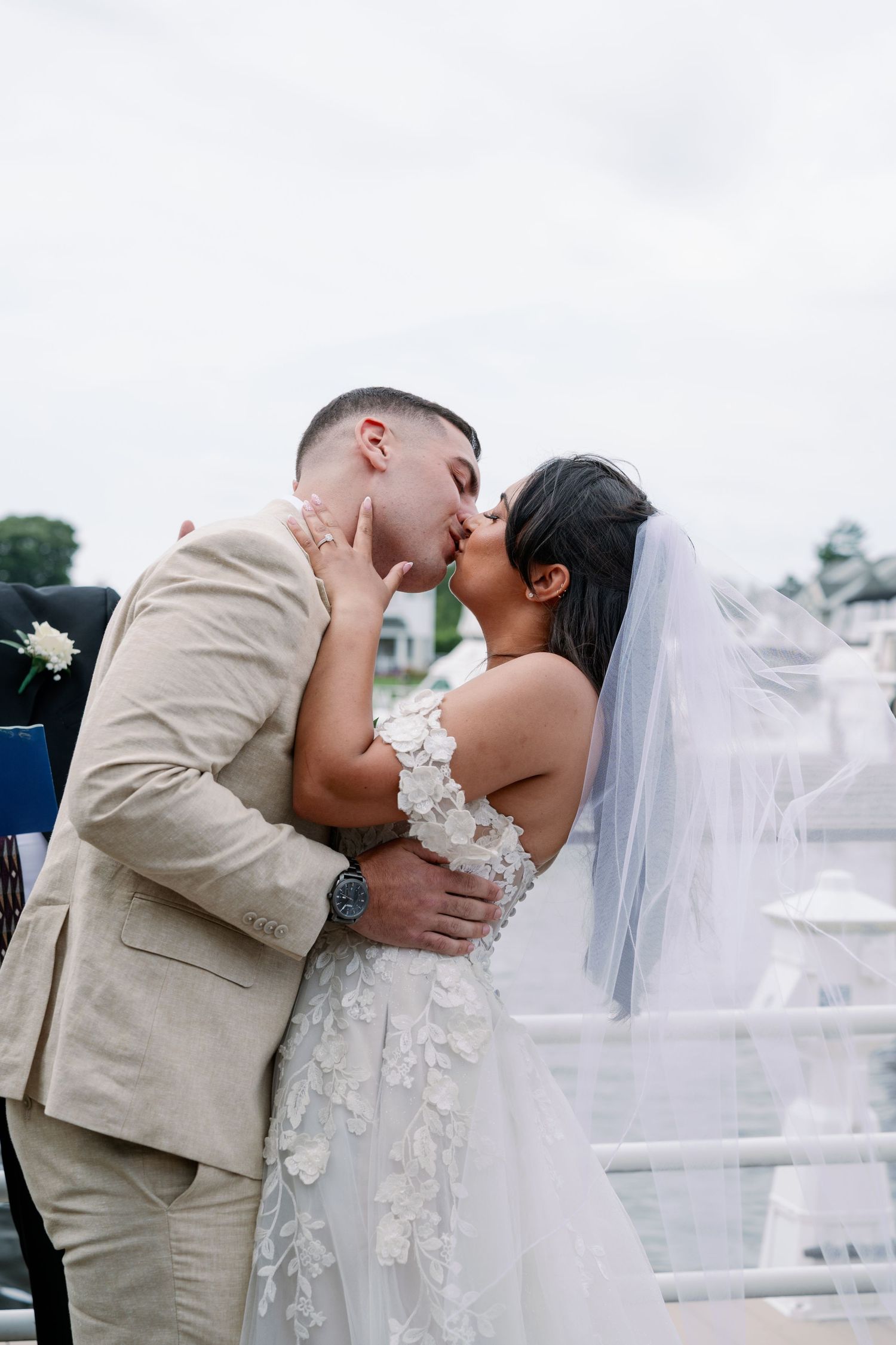 A bride in a floral lace wedding gown and veil shares a kiss with her groom in a tan suit during an outdoor waterfront ceremony.