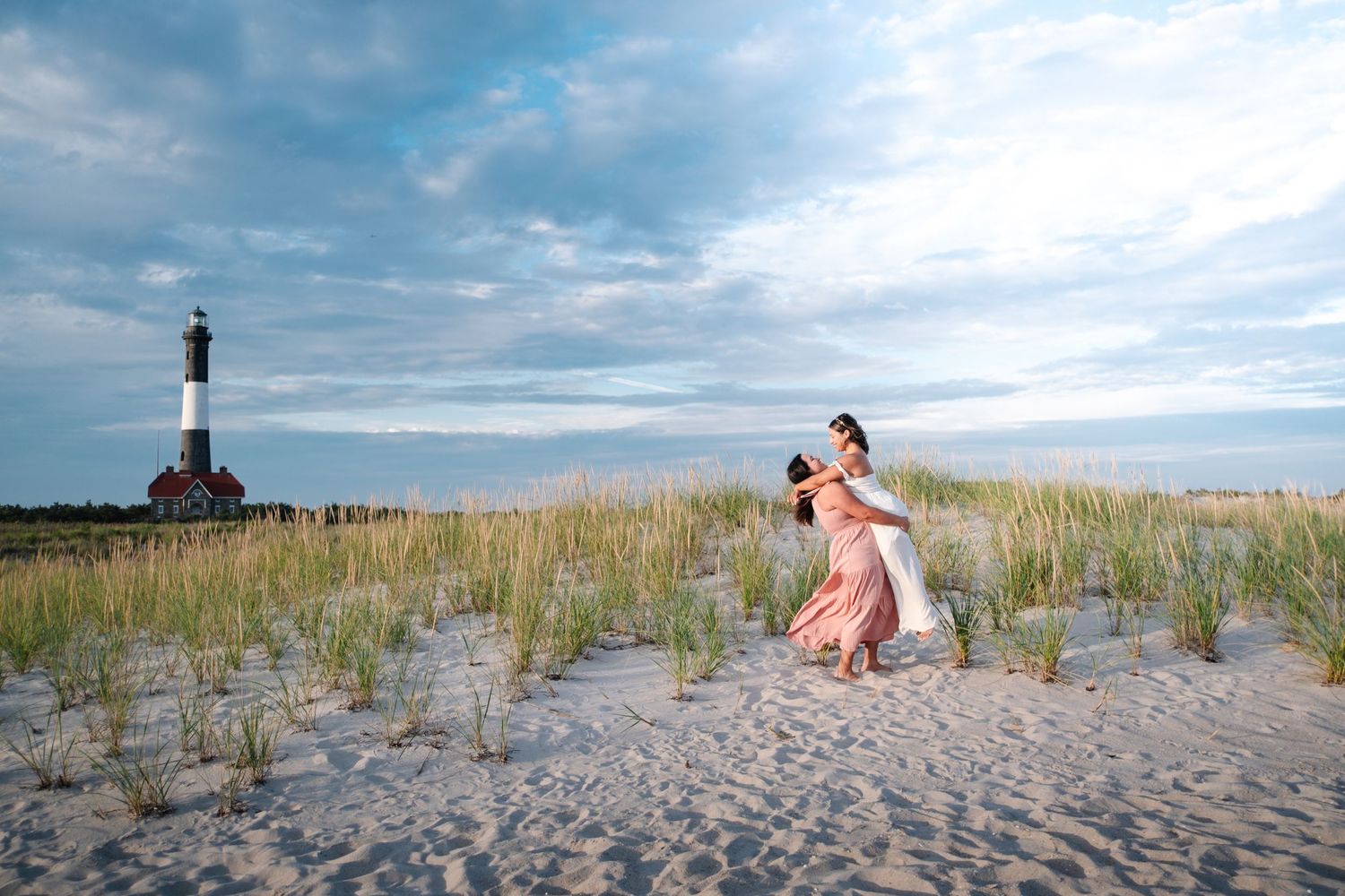 Scenic lighthouse and beach dunes with tall grass against a cloudy sky.