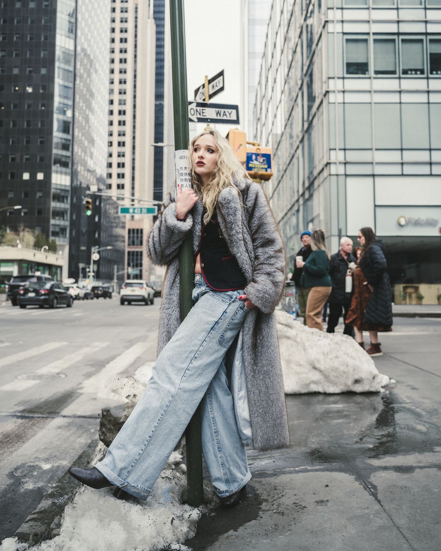 Blonde woman in a long gray fur coat and baggy light-wash jeans leans on a street pole beside a one way sign on a snowy city corner.
