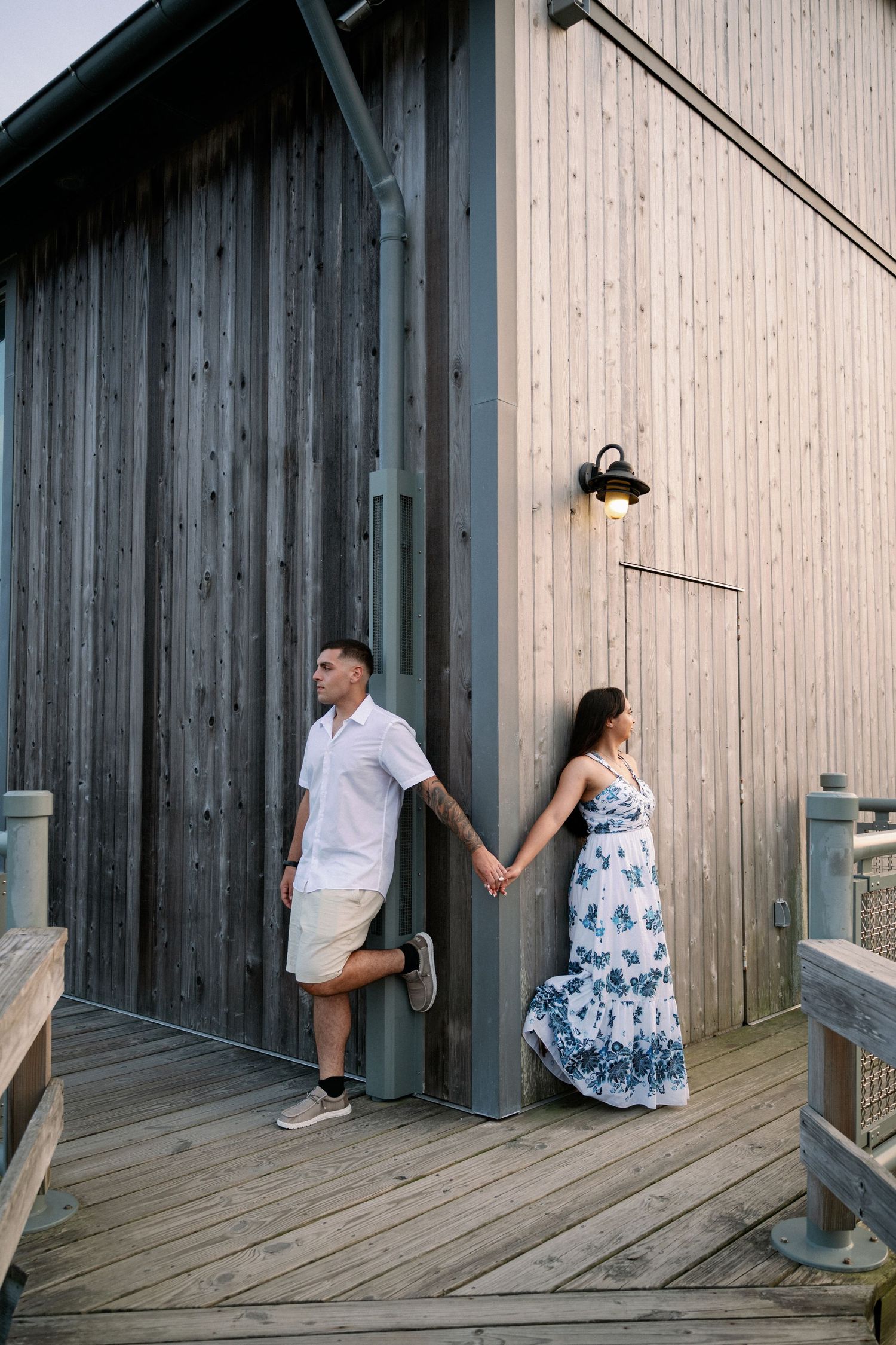 Couple holding hands on a wooden boardwalk beside a rustic building, man in white shirt and woman in floral dress.