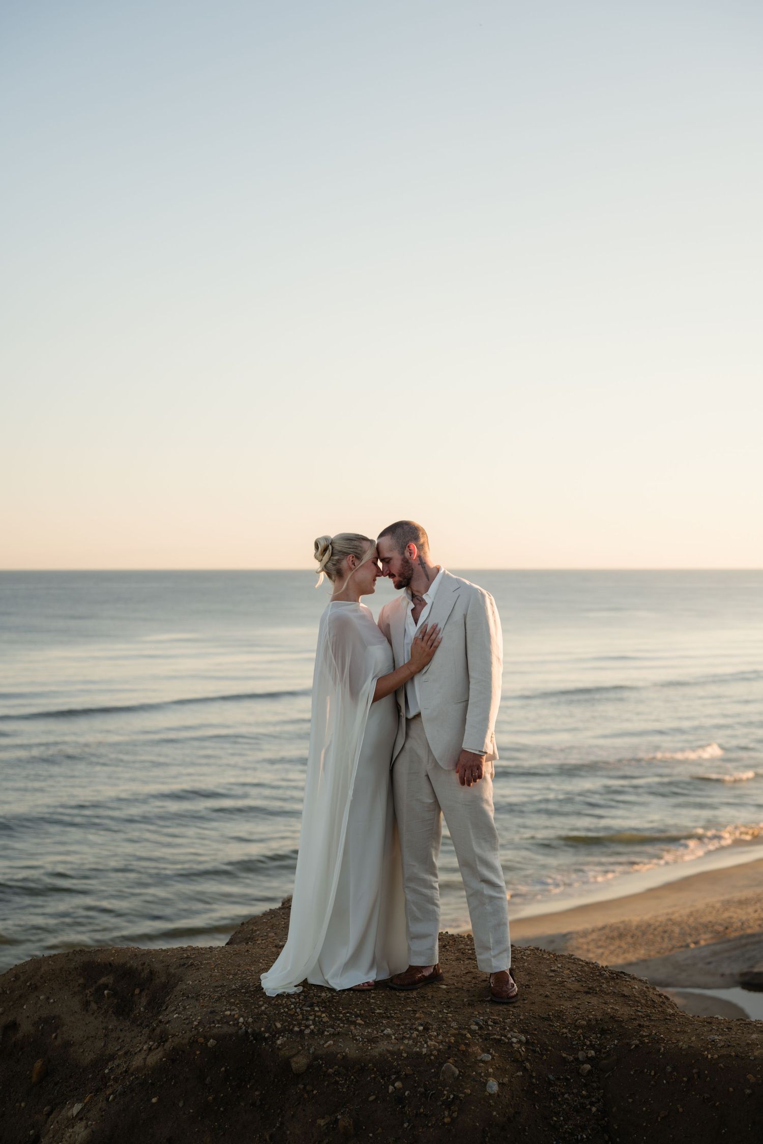 Bride in a flowing white cape gown and groom in a cream suit embrace on a coastal cliff overlooking the ocean at sunset.