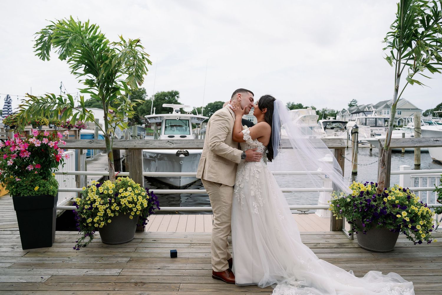 Bride in a flowing white gown and veil kisses a groom in a tan suit on a waterfront dock lined with flower pots and boats.