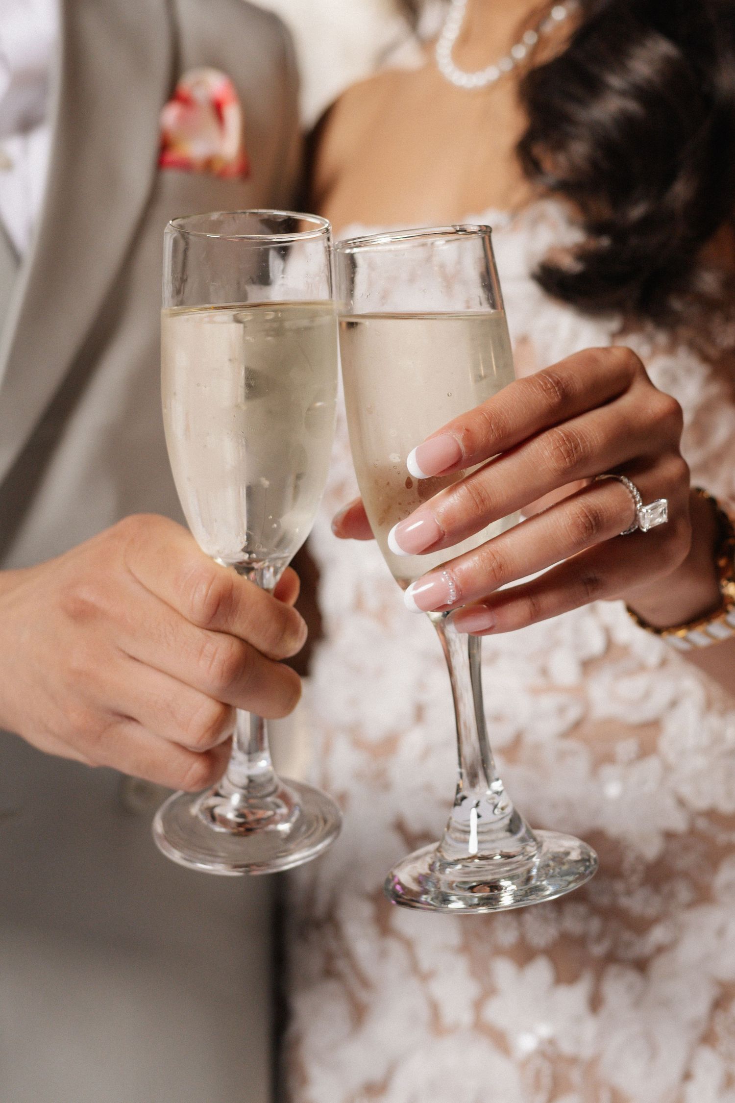 Bride and groom toast with champagne flutes at their wedding, her hand showing a sparkling diamond ring and a lace gown.